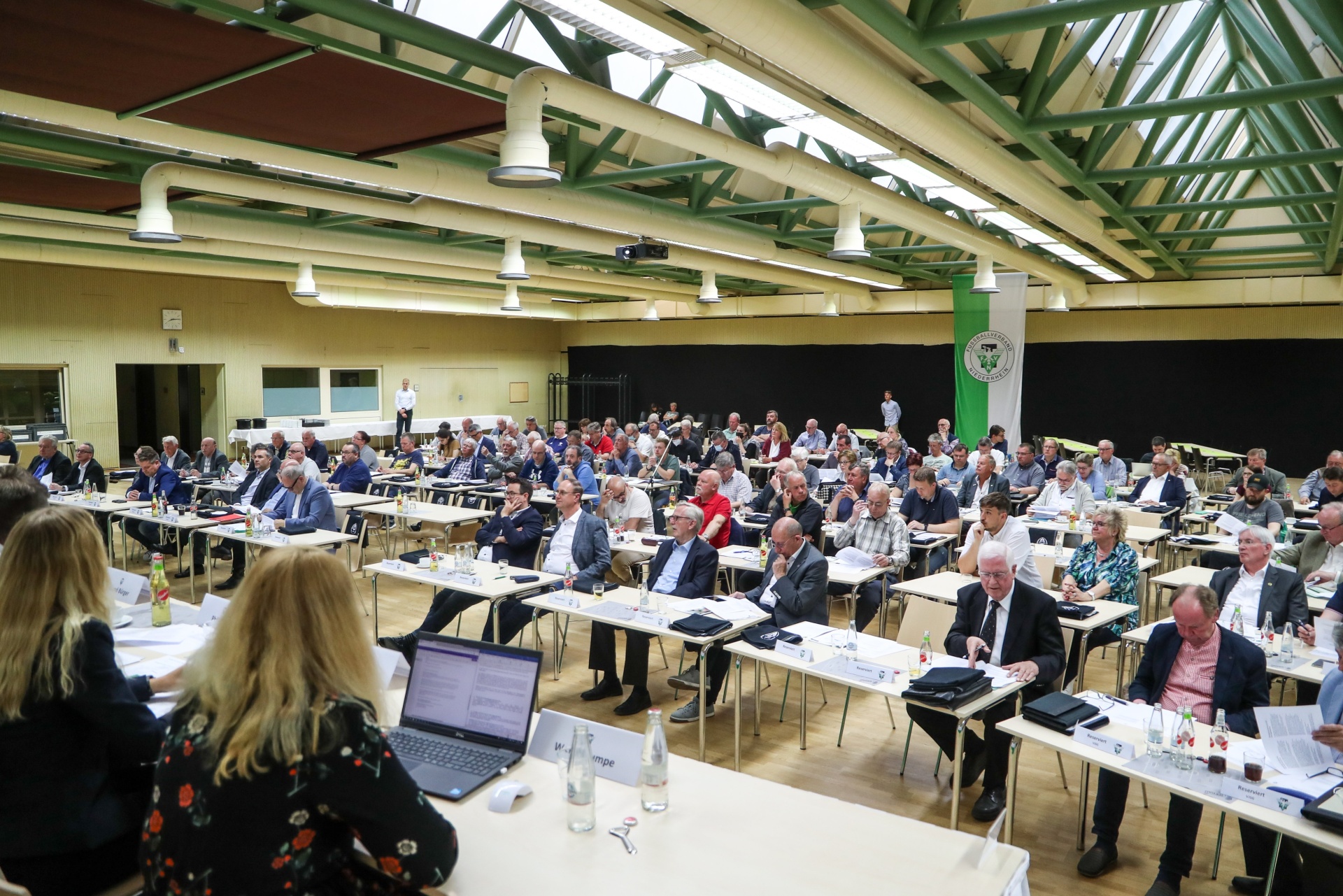 [{"clang_id":1,"value":"Großer Konferenzraum mit vielen an Tischen sitzenden Männern in Anzügen. Im Vordergrund Frau mit Laptop, Tische mit Wasser und Notizen."}]