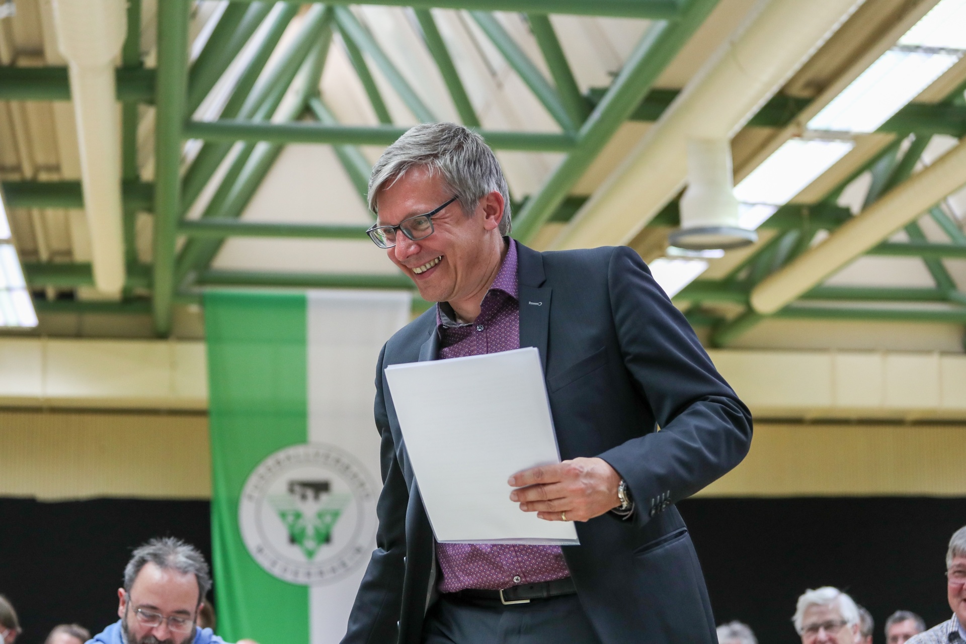 [{"clang_id":1,"value":"Mann mit Brille im Anzug liest ein Blatt Papier, vermutlich während einer Rede in einer großen Halle. Im Hintergrund Personen und eine grüne Flagge."}]
