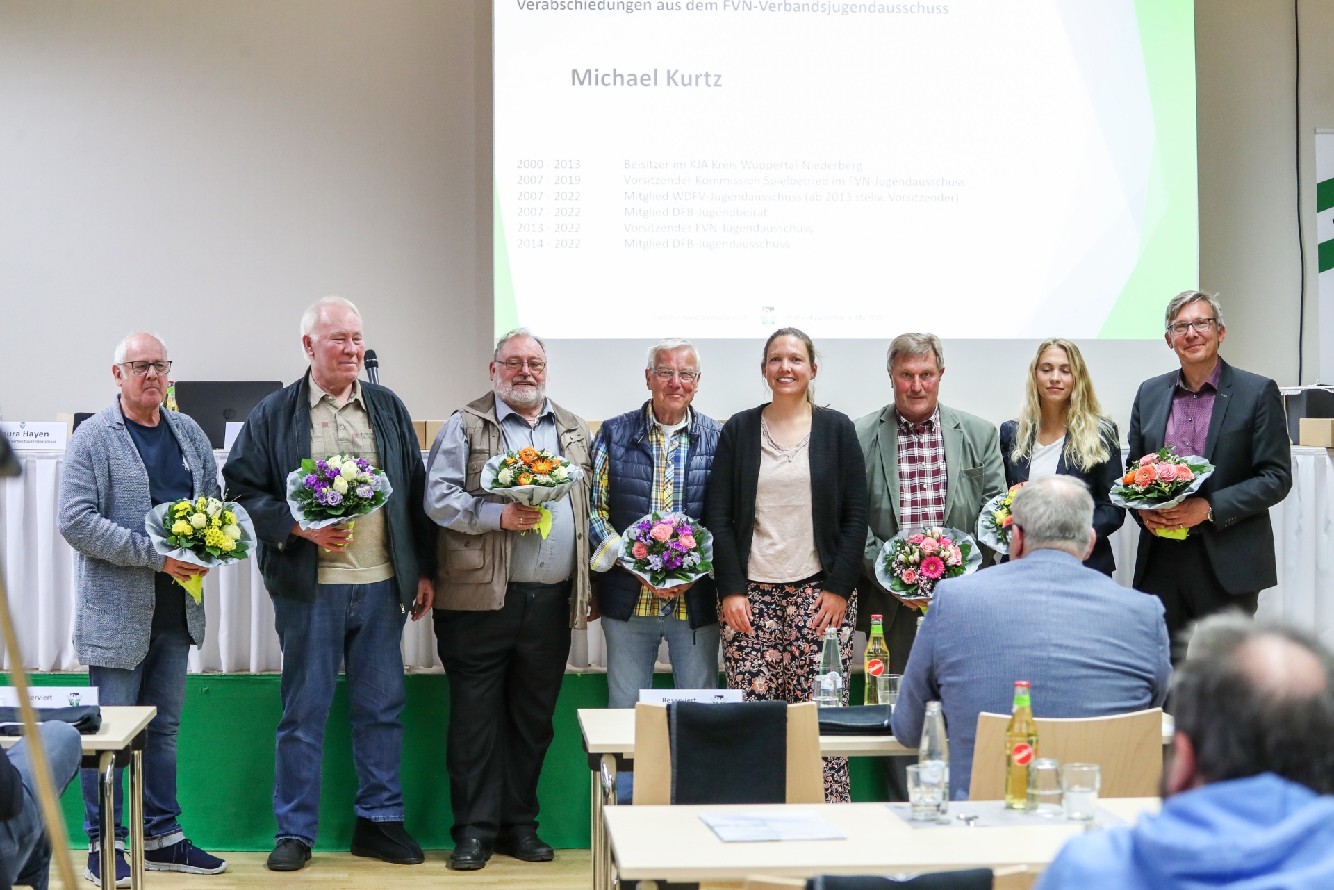 [{"clang_id":1,"value":"Gruppe von acht Personen auf einer Bühne, die Blumensträuße entgegennehmen. Im Hintergrund eine Präsentation mit dem Namen \"Michael Kurtz\" und Jahreszahlen."}]