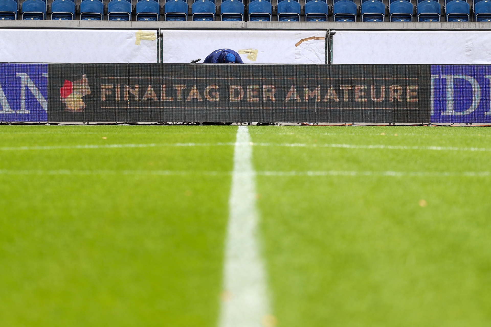 [{"clang_id":1,"value":"Fußballfeld mit weißer Mittellinie. Im Hintergrund ein Werbebanner mit Text in deutscher Sprache: \"Finaltag der Amateure\". Leere Zuschauertribüne."}]