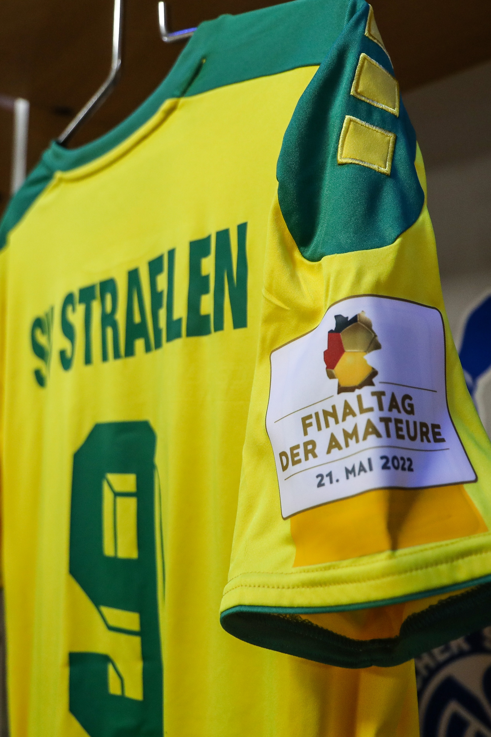 [{"clang_id":1,"value":"Gelbes Fußballtrikot mit grünem Kragen und der Aufschrift \"STRAFLEN\" sowie der Nummer 9. Auf der Brust ein Patch mit der Aufschrift \"FINALTAG DER AMATEURE 21. MAI 2022\"."}]