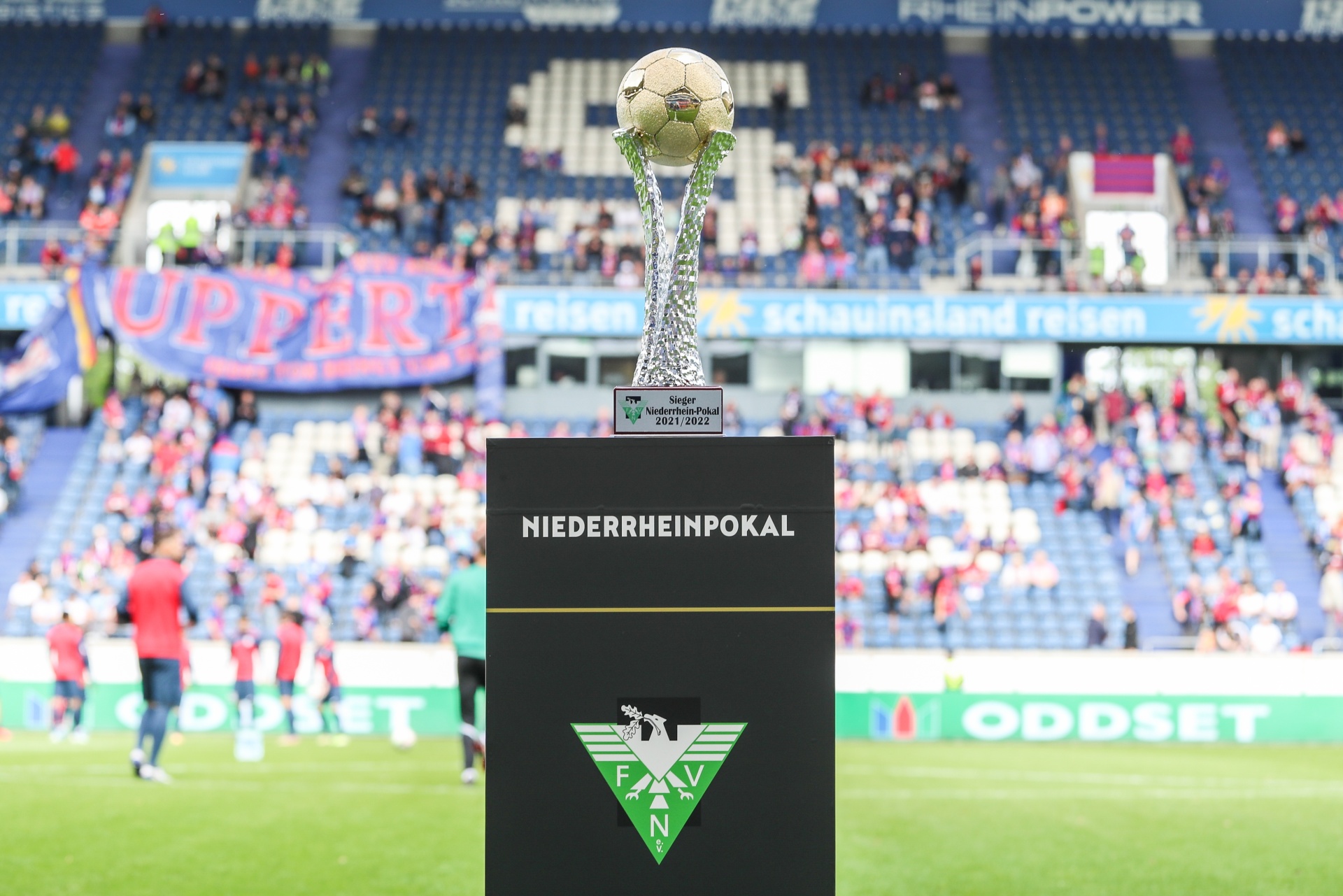 [{"clang_id":1,"value":"Fußballstadion: Pokal auf grünem Sockel im Vordergrund, dahinter Fans auf den Tribünen. Im Hintergrund Werbebanner."}]