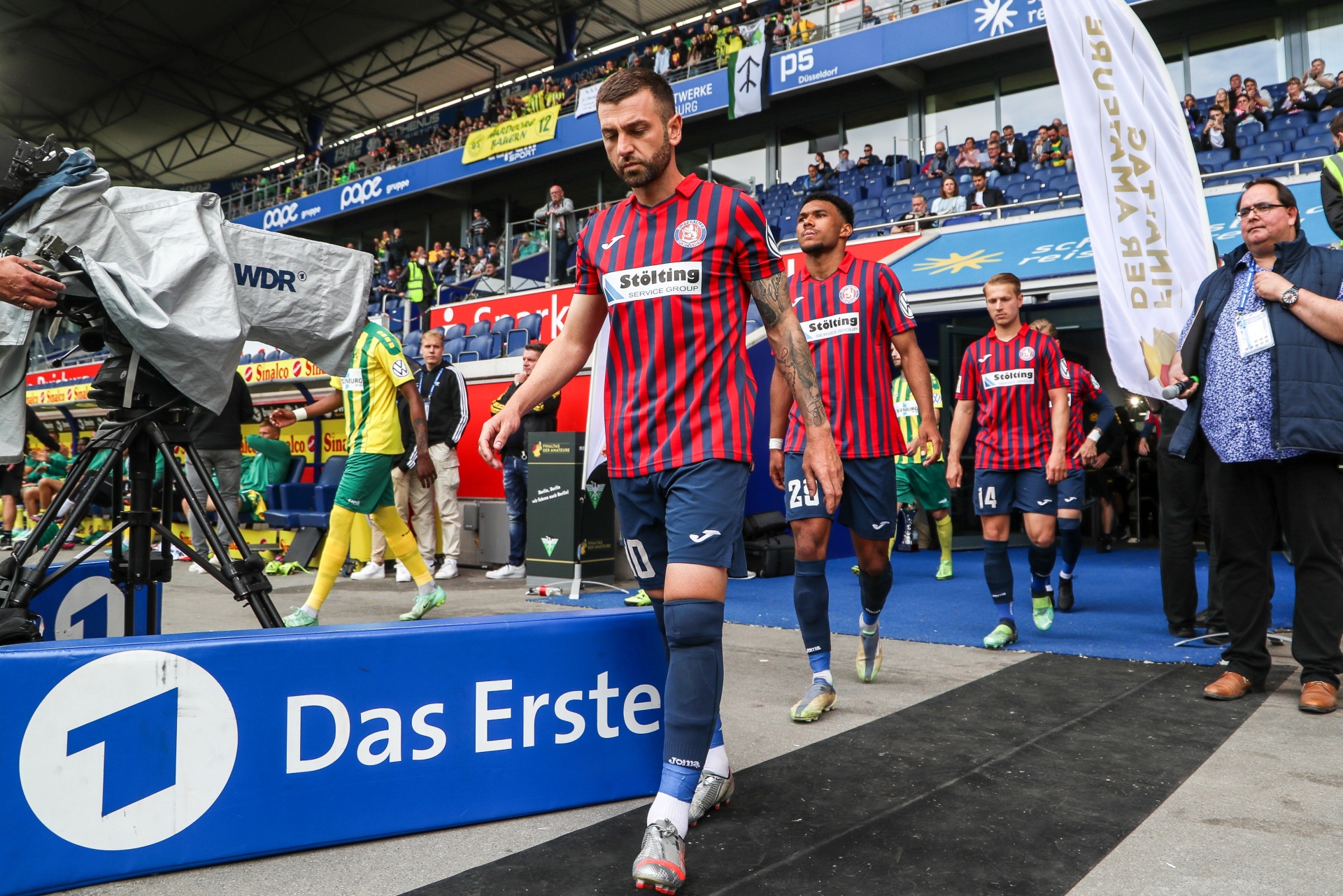 [{"clang_id":1,"value":"Fußballspieler in roten und schwarzen Trikots betreten einen Stadiontunnel. Im Vordergrund ein Banner mit dem Logo \"Das Erste\". Zuschauerreihen im Hintergrund."}]