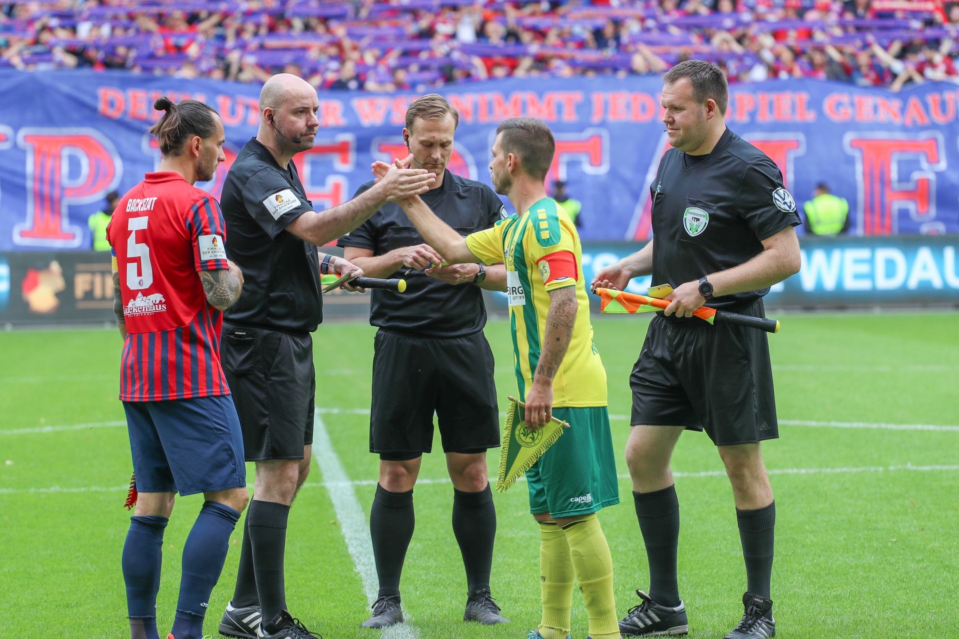 [{"clang_id":1,"value":"Fußballspiel: Zwei Spieler (rot/schwarz, grün/gelb) diskutieren mit zwei Schiedsrichtern. Im Hintergrund Zuschauer und Werbebanner."}]