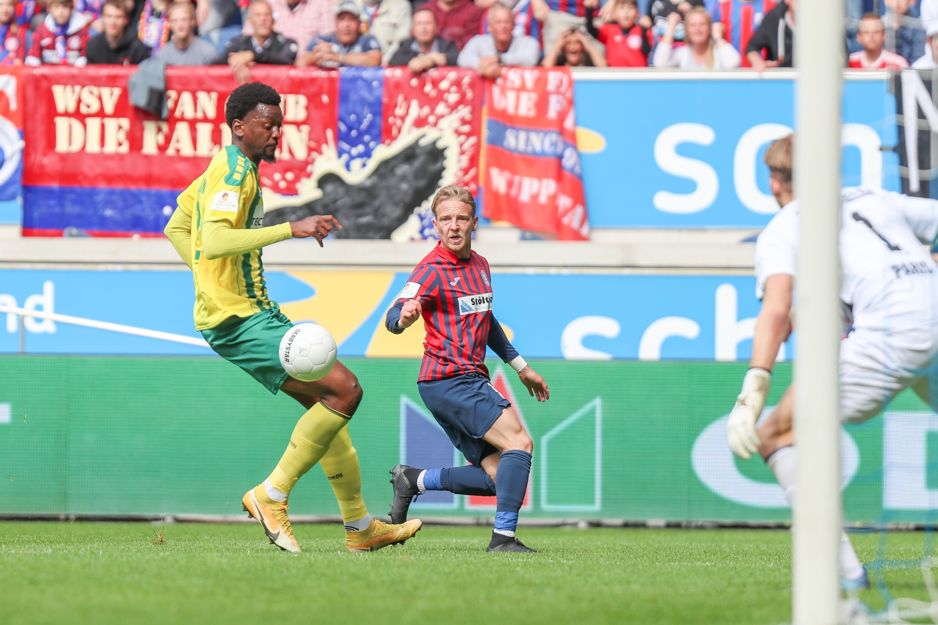 [{"clang_id":1,"value":"Fußballspiel: Zwei Spieler verfolgen Ball. Im Hintergrund Banner und Zuschauer. Torwart steht im Tor. Rasenplatz."}]