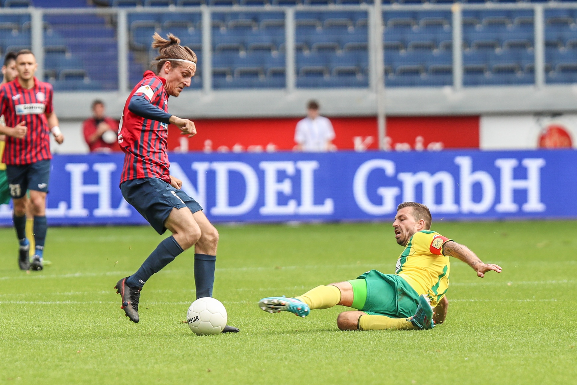 [{"clang_id":1,"value":"Fußballspiel: Spieler in Rot/Schwarz dribbelt an Gegenspieler in Grün/Gelb vorbei. Im Hintergrund Zuschauerränge und Werbebanner."}]