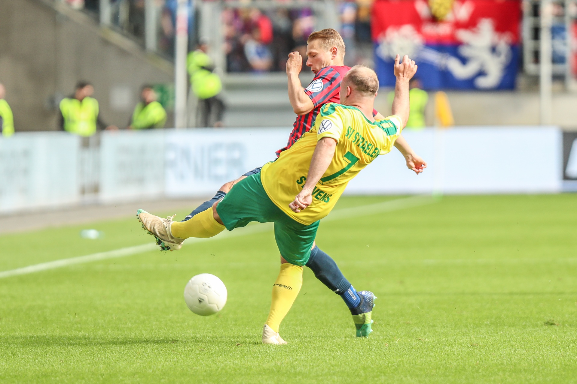 [{"clang_id":1,"value":"Zwei Fußballspieler kämpfen um den Ball auf grünem Rasen. Ein Spieler in grün-gelbem Trikot versucht, den Ball zu treten, während ein Gegner in blau-rotem Trikot ihn blockt. Im Hintergrund Zuschauer."}]