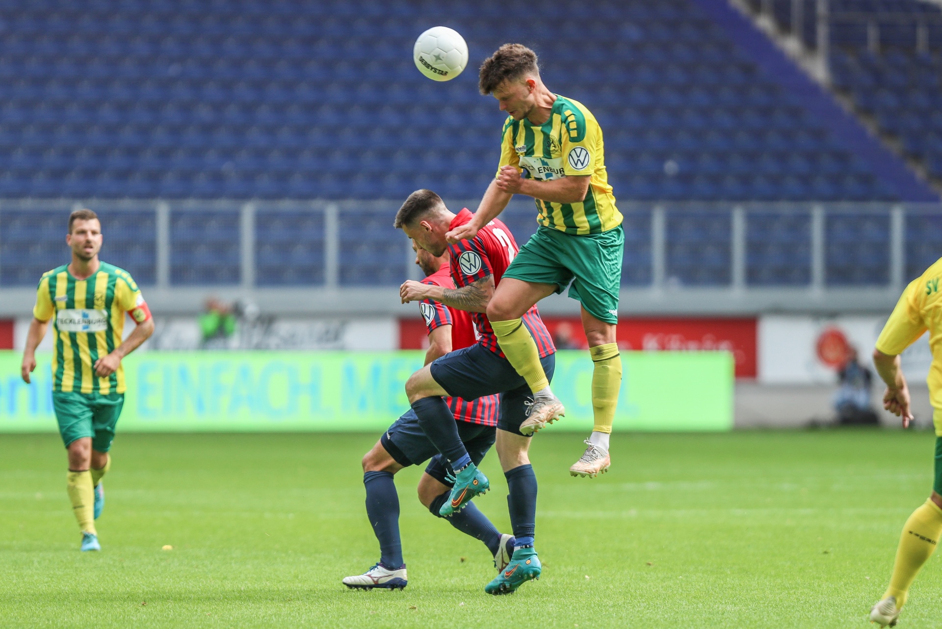 [{"clang_id":1,"value":"Zwei Fußballspieler springen um einen Ball. Ein Spieler in grün-gelbem Trikot über einem in rot-schwarz. Im Hintergrund weitere Spieler und Zuschauerränge."}]