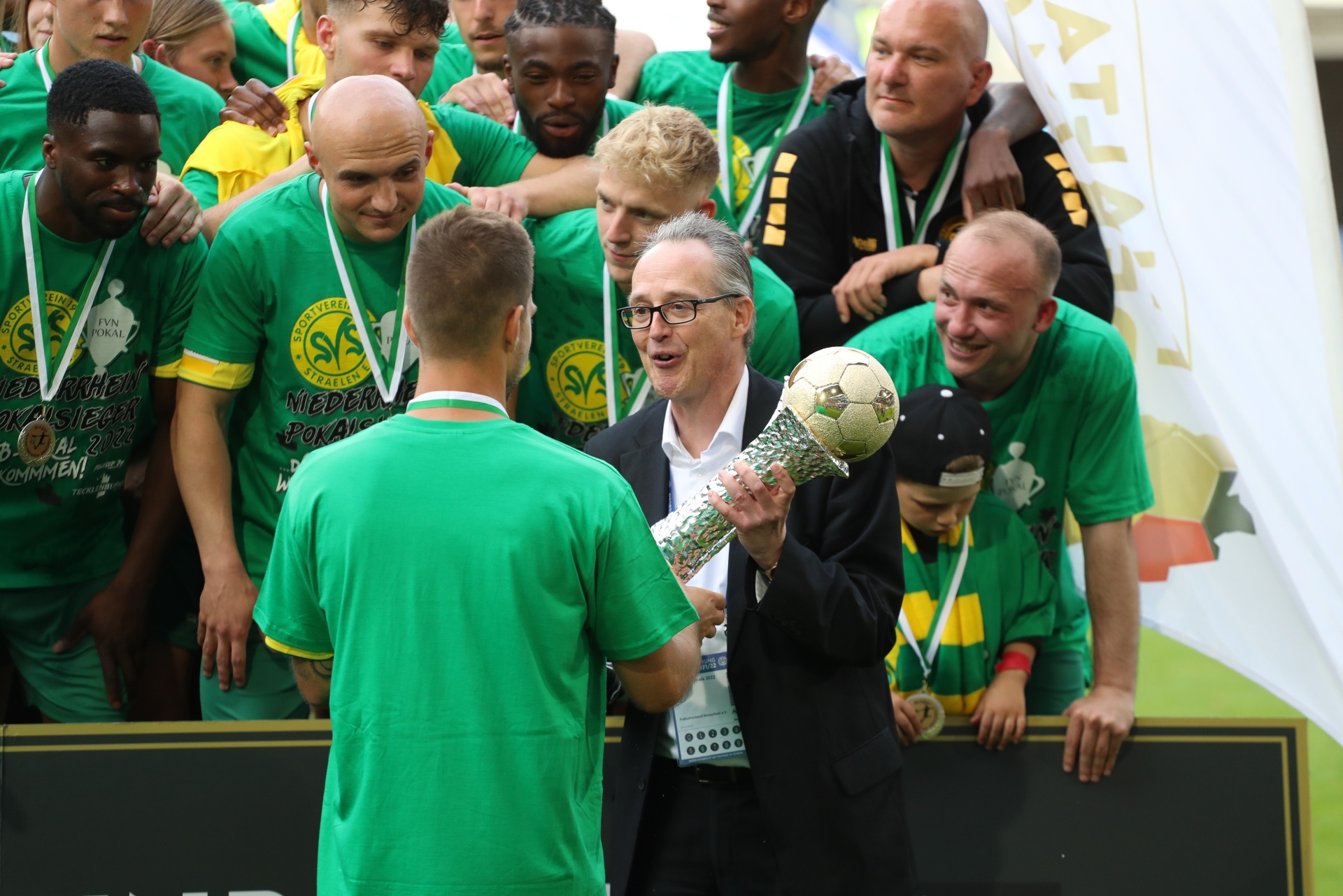 [{"clang_id":1,"value":"Fußballmannschaft in grünen Trikots feiert Sieg. Ein Mann überreicht dem Kapitän einen goldenen Pokal. Im Hintergrund weitere Spieler und Betreuer."}]