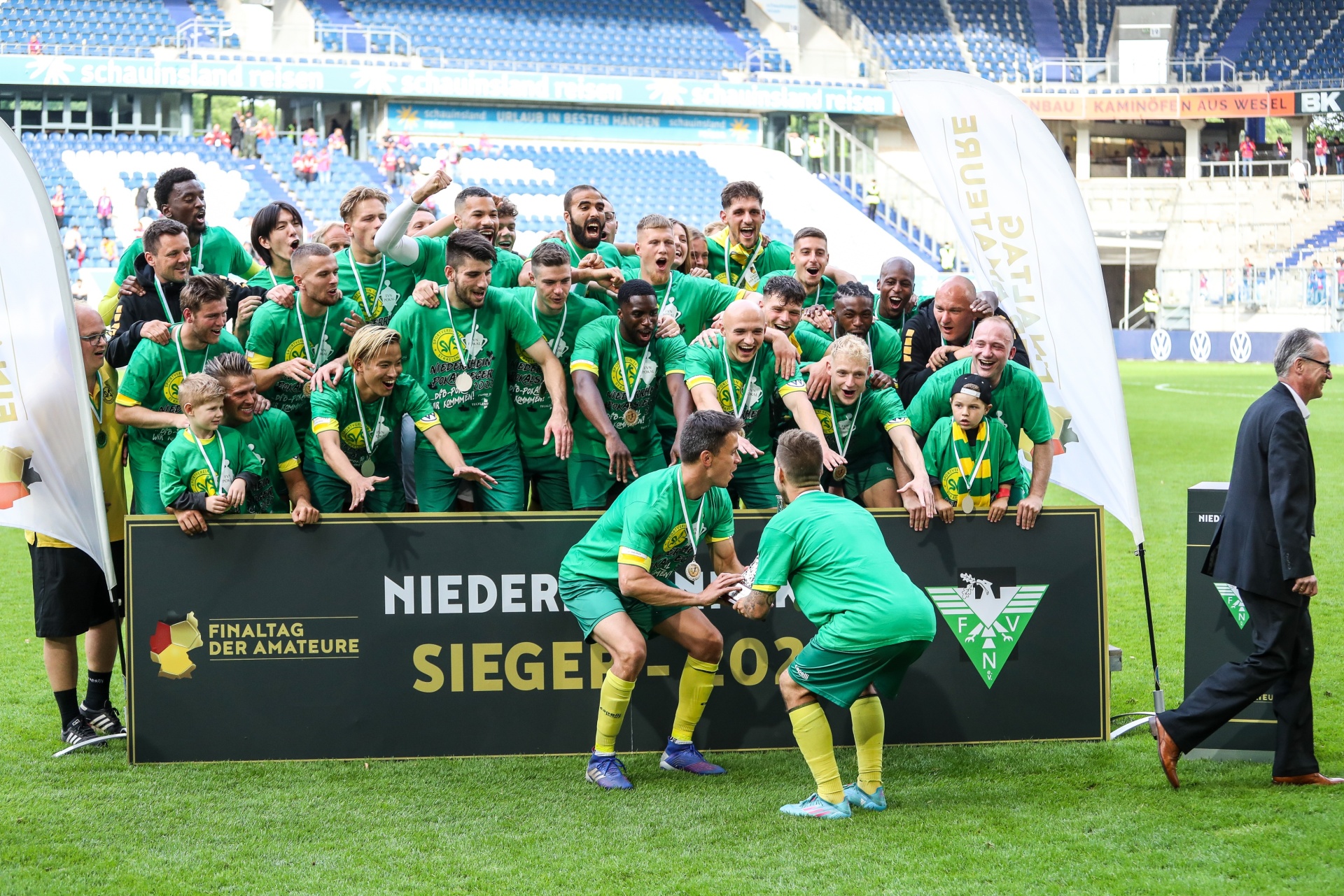 [{"clang_id":1,"value":"Fußballmannschaft in grünen Trikots jubelt vor einer schwarzen Tafel mit der Aufschrift \"NIEDER SIEGER\". Spieler tragen Medaillen, im Hintergrund Stadiontribüne."}]
