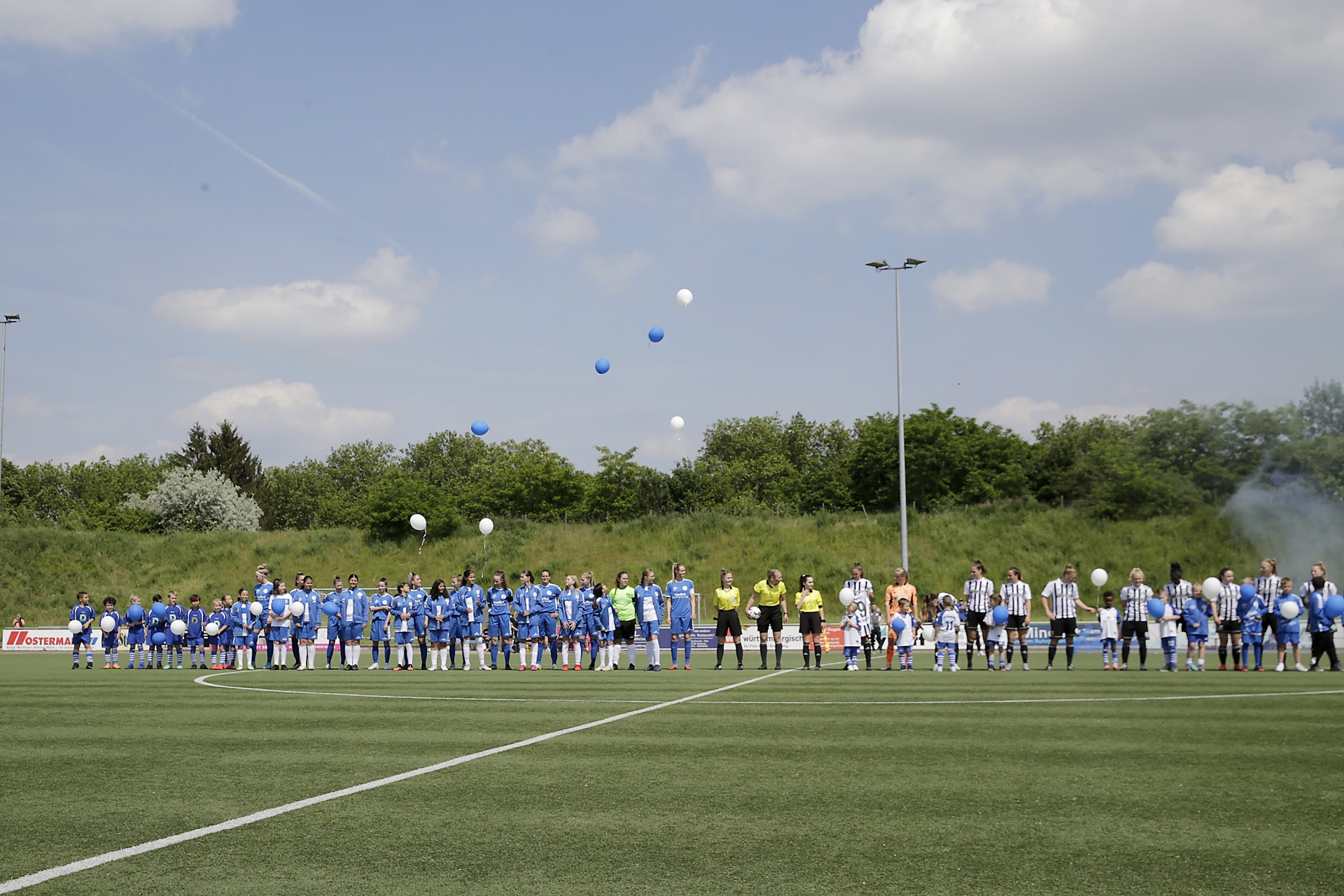 [{"clang_id":1,"value":"Fußballspiel: Zwei Mannschaften stehen sich auf grünem Rasen gegenüber. Spieler tragen blaue bzw. schwarz-weiße Trikots. Im Hintergrund Bäume und ein blauer Himmel. Ballons steigen auf."}]