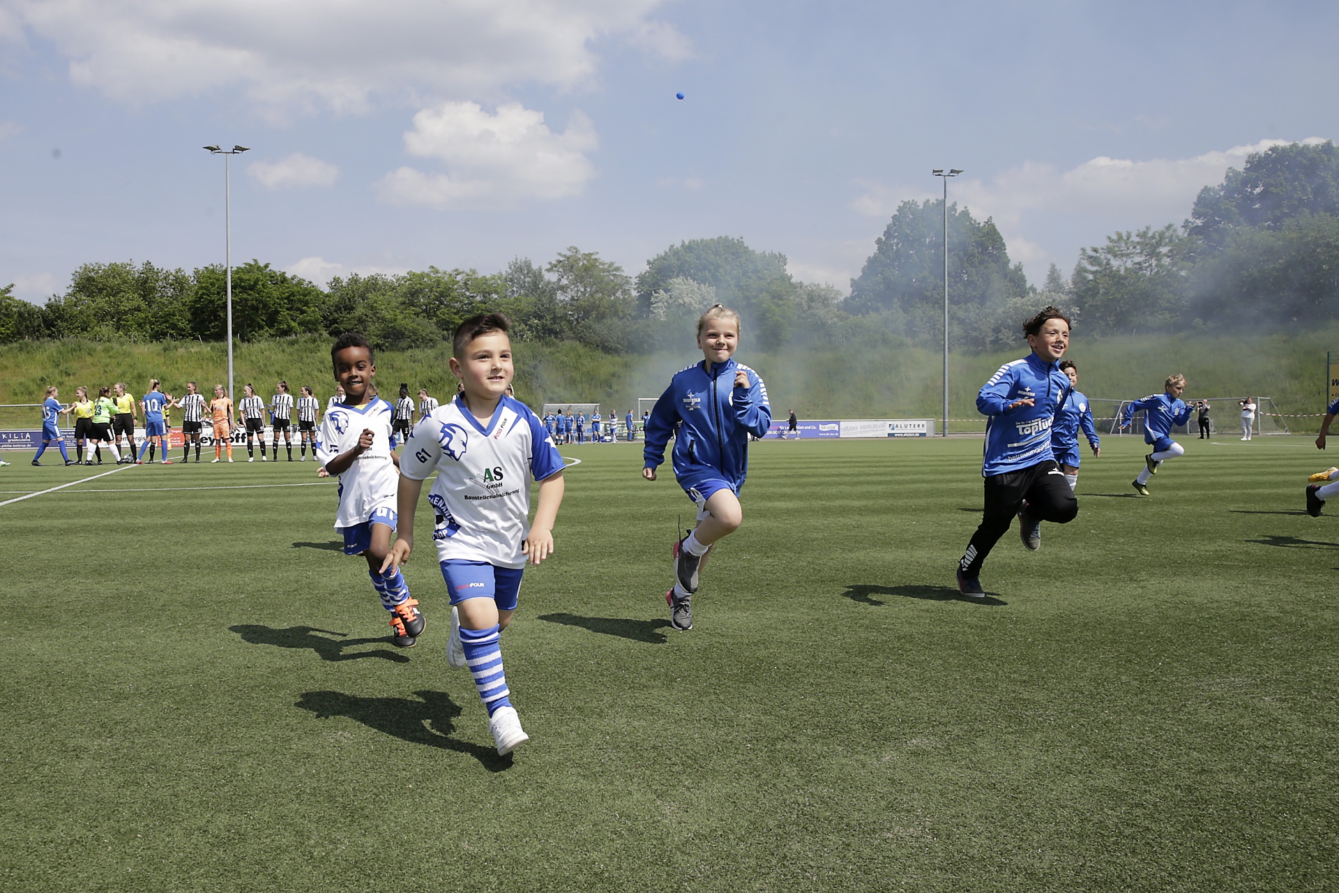 [{"clang_id":1,"value":"Fußballspiel mit Kindern auf Kunstrasenfeld. Vier Läufer im Vordergrund, weitere Spieler und Zuschauer im Hintergrund unter blauem Himmel."}]