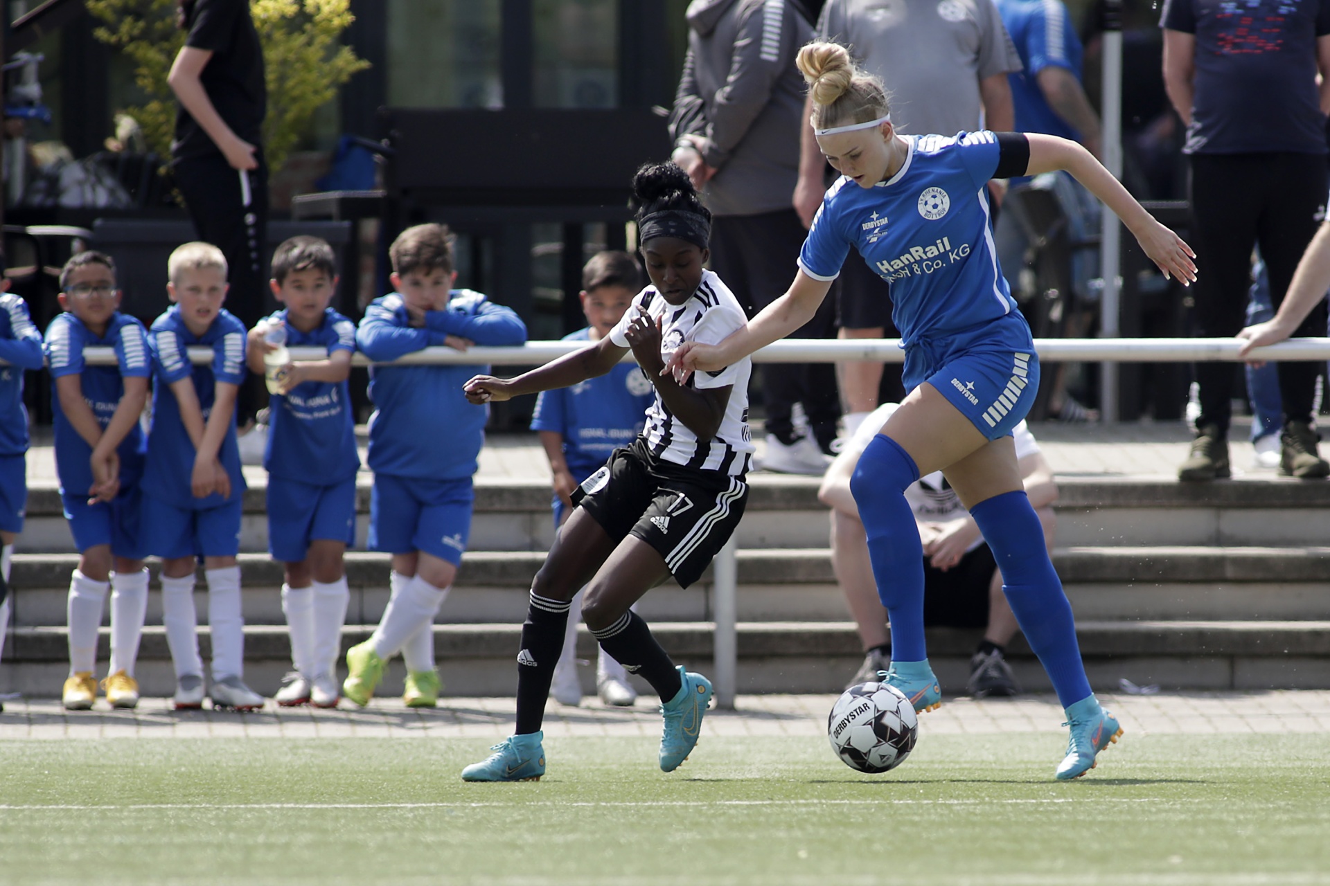 [{"clang_id":1,"value":"Fußballspiel: Zwei Spielerinnen kämpfen um den Ball. Im Hintergrund Zuschauer und weitere Spieler. Rasenplatz und Tribüne."}]