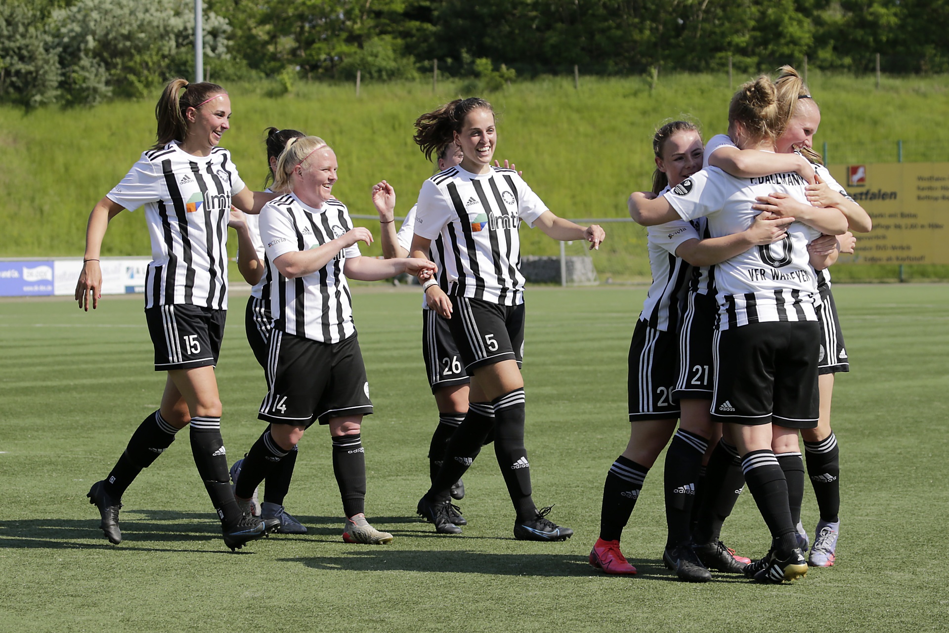 [{"clang_id":1,"value":"Frauenfußballteam jubelt auf grünem Rasen. Spielerinnen in schwarz-weißen Trikots umarmen sich und rennen, einige mit erhobenen Armen. Im Hintergrund eine Bank und grüne Hügel."}]