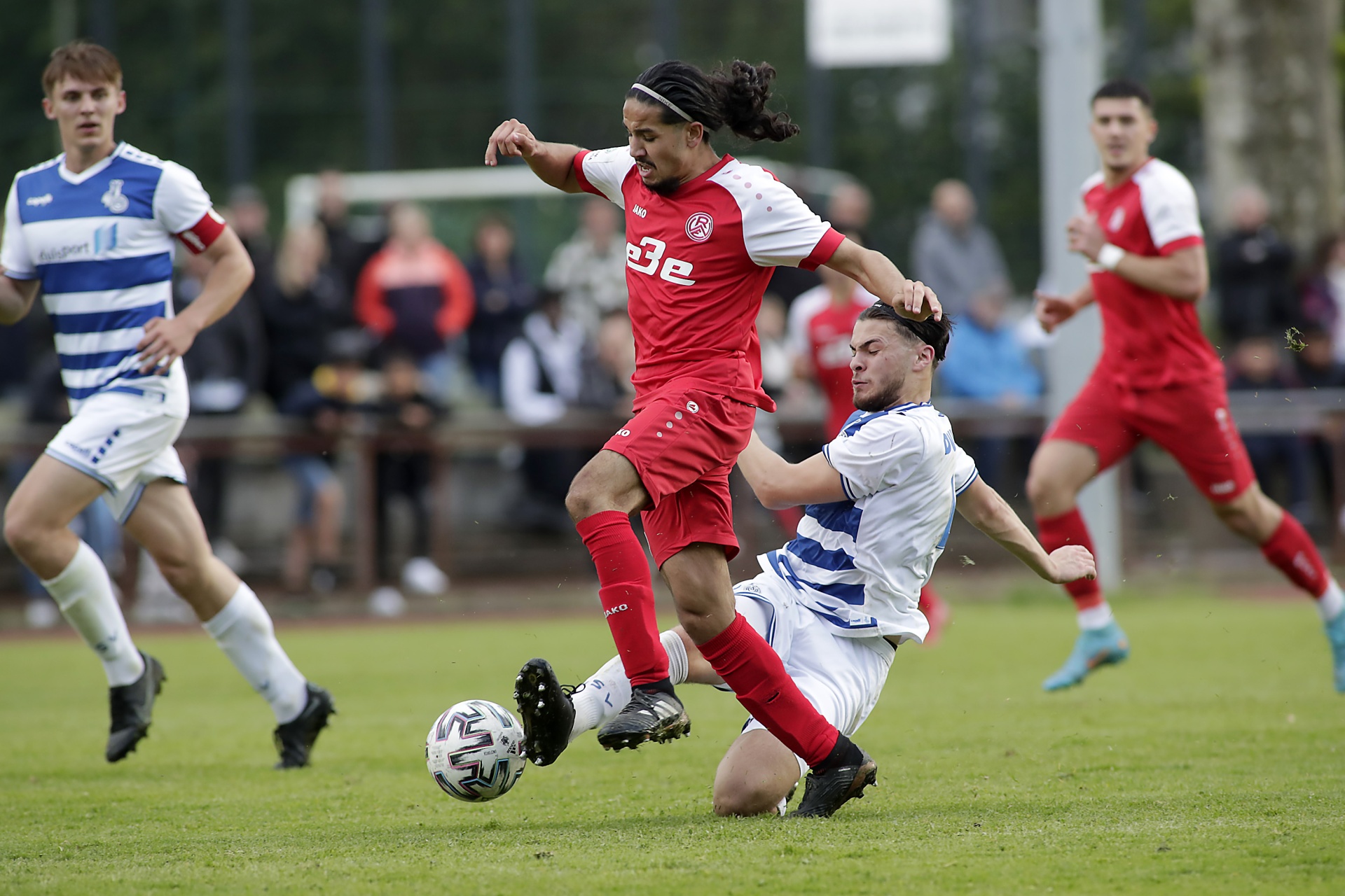 [{"clang_id":1,"value":"Fußballspiel: Spieler mit langen Haaren in rotem Trikot dribbelt am Ball, wird von Gegenspieler in blau-weißem Trikot gefoult. Im Hintergrund Zuschauer und weitere Spieler."}]