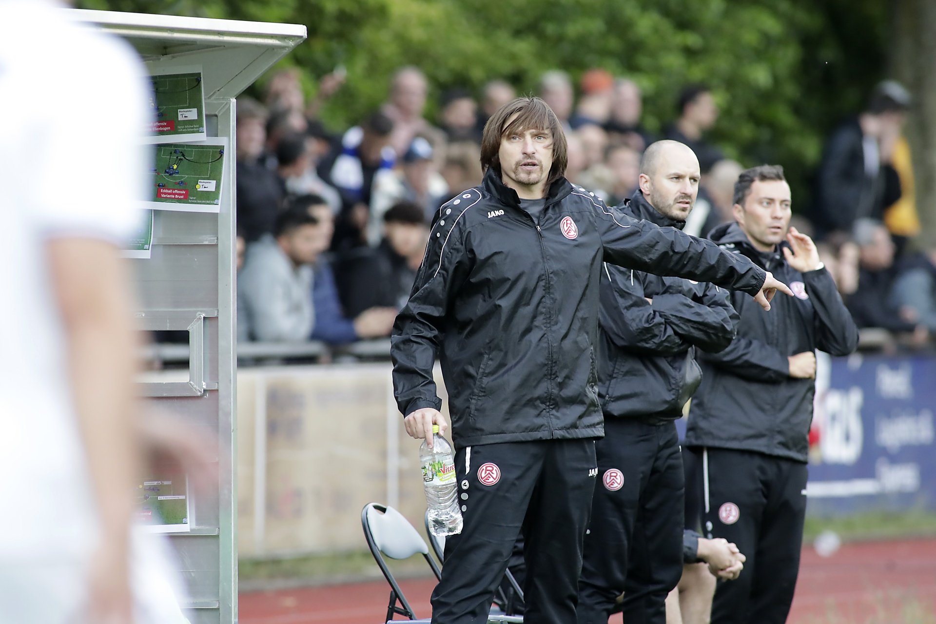 [{"clang_id":1,"value":"Fußballtrainer mit Team am Spielfeldrand. Trainer hält Wasserflasche, alle tragen Trainingsanzüge mit rotem Logo. Im Hintergrund Zuschauer und ein Stadiondach."}]
