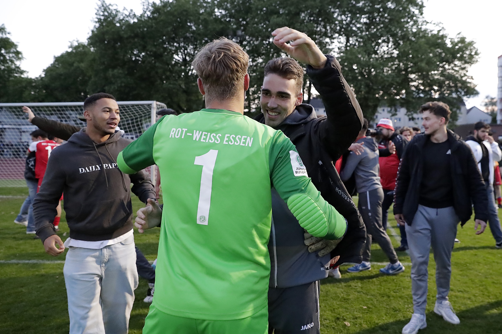 [{"clang_id":1,"value":"Fußballspieler im grünen Trikot mit Rückennummer 1 wird von zwei Personen umarmt. Im Hintergrund weitere Fußballspieler und eine Sportanlage."}]