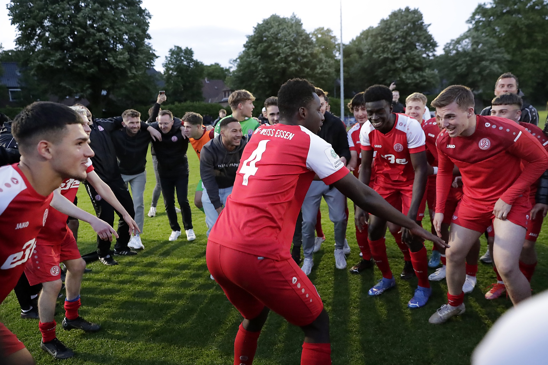 [{"clang_id":1,"value":"Fußballspieler in roten Trikots feiern im Kreis auf einem Fußballplatz. Einige stehen im Hintergrund, andere umarmen sich. Bäume im Hintergrund."}]