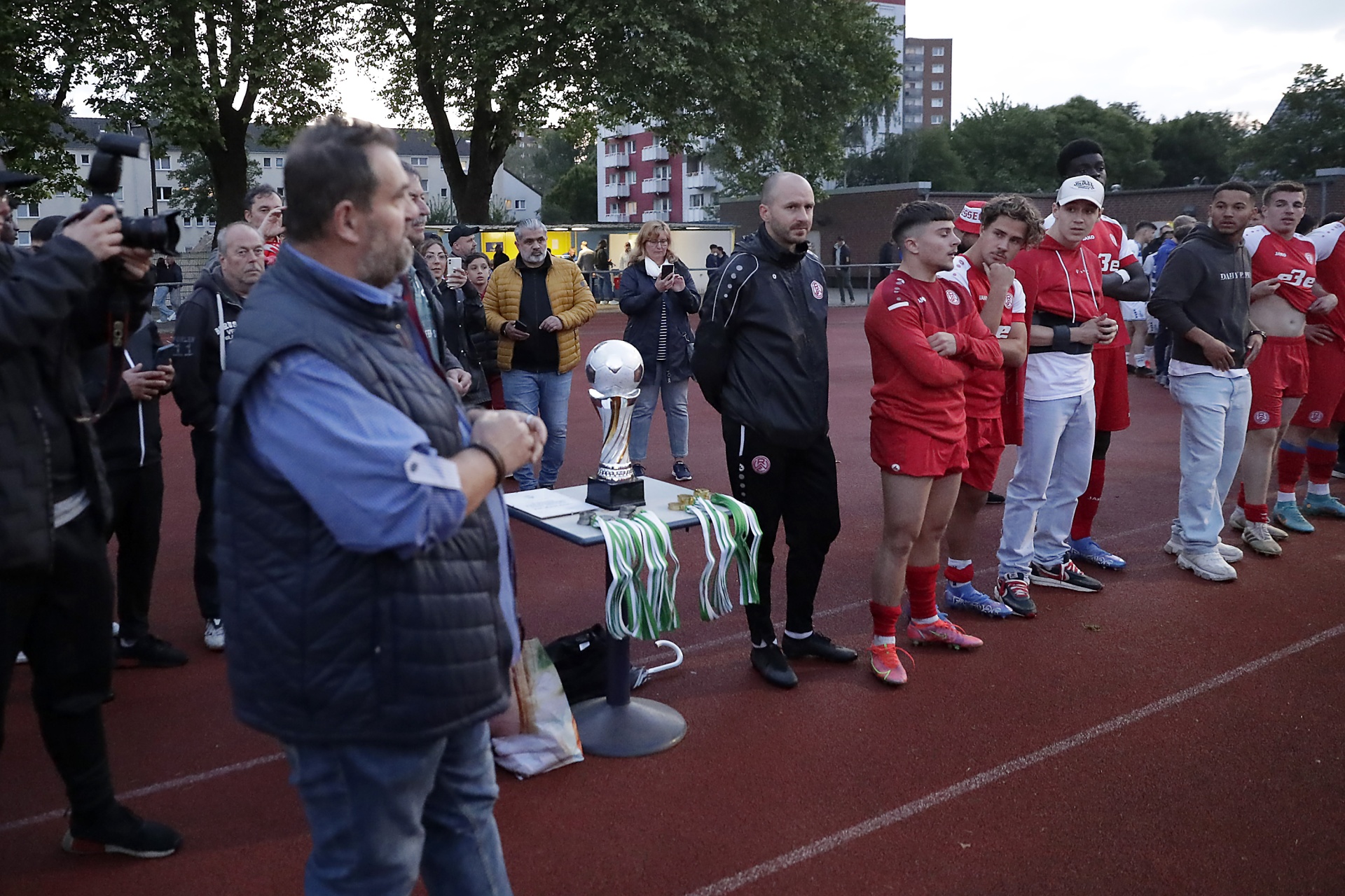 [{"clang_id":1,"value":"Männer stehen auf einem roten Sportplatz um eine Trophäe herum. Einige tragen rote Sportkleidung, andere Alltagskleidung. Im Hintergrund Gebäude und Bäume."}]