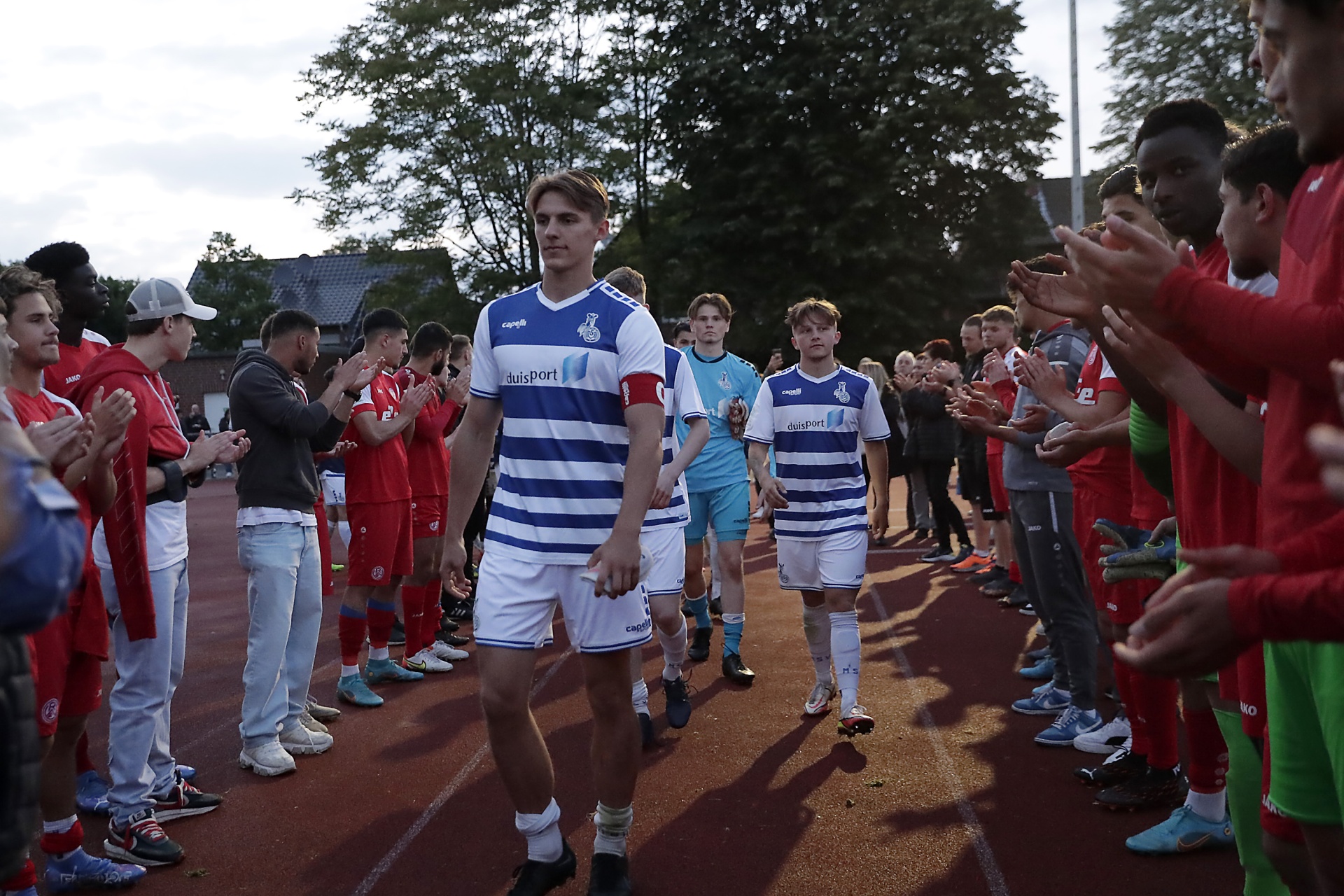 [{"clang_id":1,"value":"Fußballspieler in gestreiften Trikots betreten einen Laufbahn, umgeben von applaudierenden Zuschauern in roten und grünen Trainingsanzügen."}]
