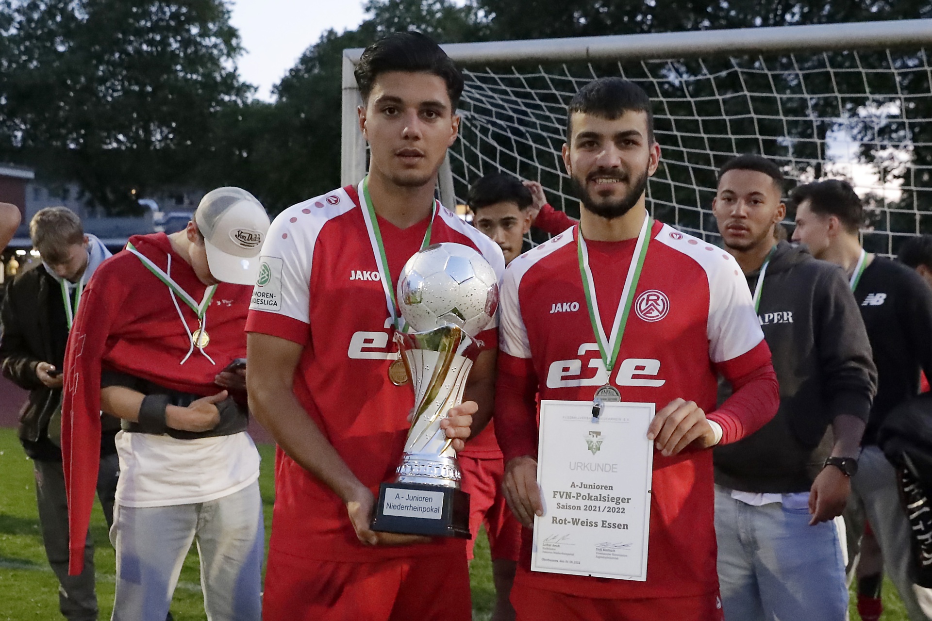 [{"clang_id":1,"value":"Fußballspieler in rot-weißen Trikots mit Pokal und Urkunde nach dem Gewinn eines Wettbewerbs. Im Hintergrund Fußballtor und Zuschauer."}]