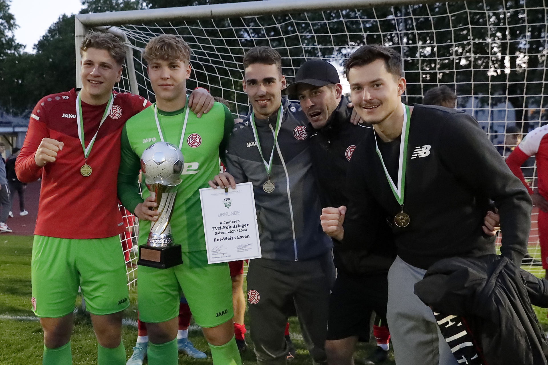 [{"clang_id":1,"value":"Fünf junge Männer in Sportkleidung und Anzug stehen auf einem Fußballfeld mit Pokal und Medaillen. Im Hintergrund ein Fußballtor."}]