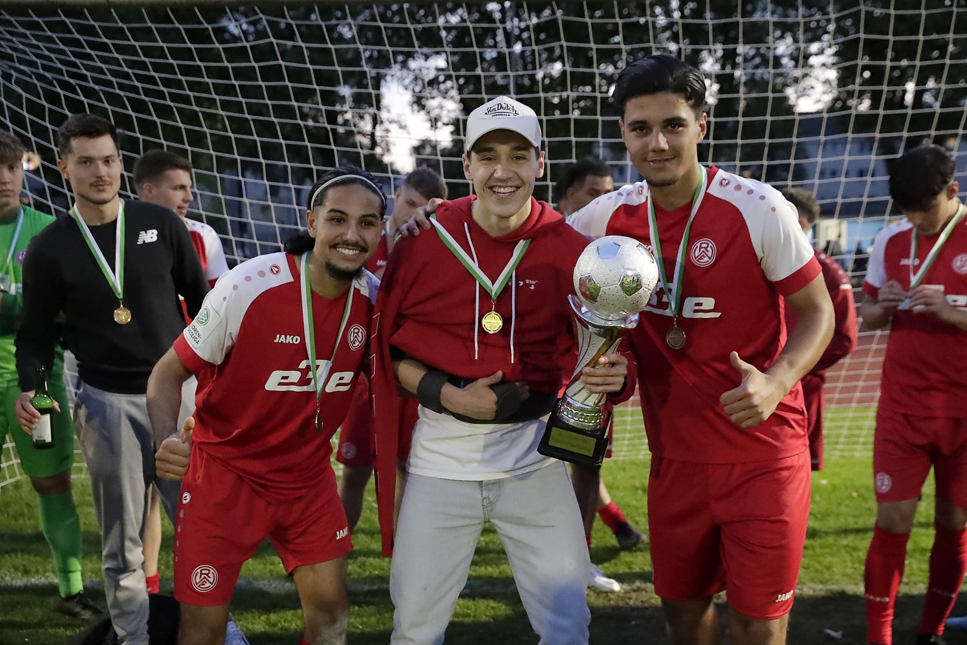 [{"clang_id":1,"value":"Fußballteam in roten Trikots mit Pokal und Medaillen nach einem gewonnenen Spiel. Ein Mann in weißem Hemd steht mittig mit dem Pokal. Hintergrund: Fußballtor und jubelnde Teammitglieder."}]