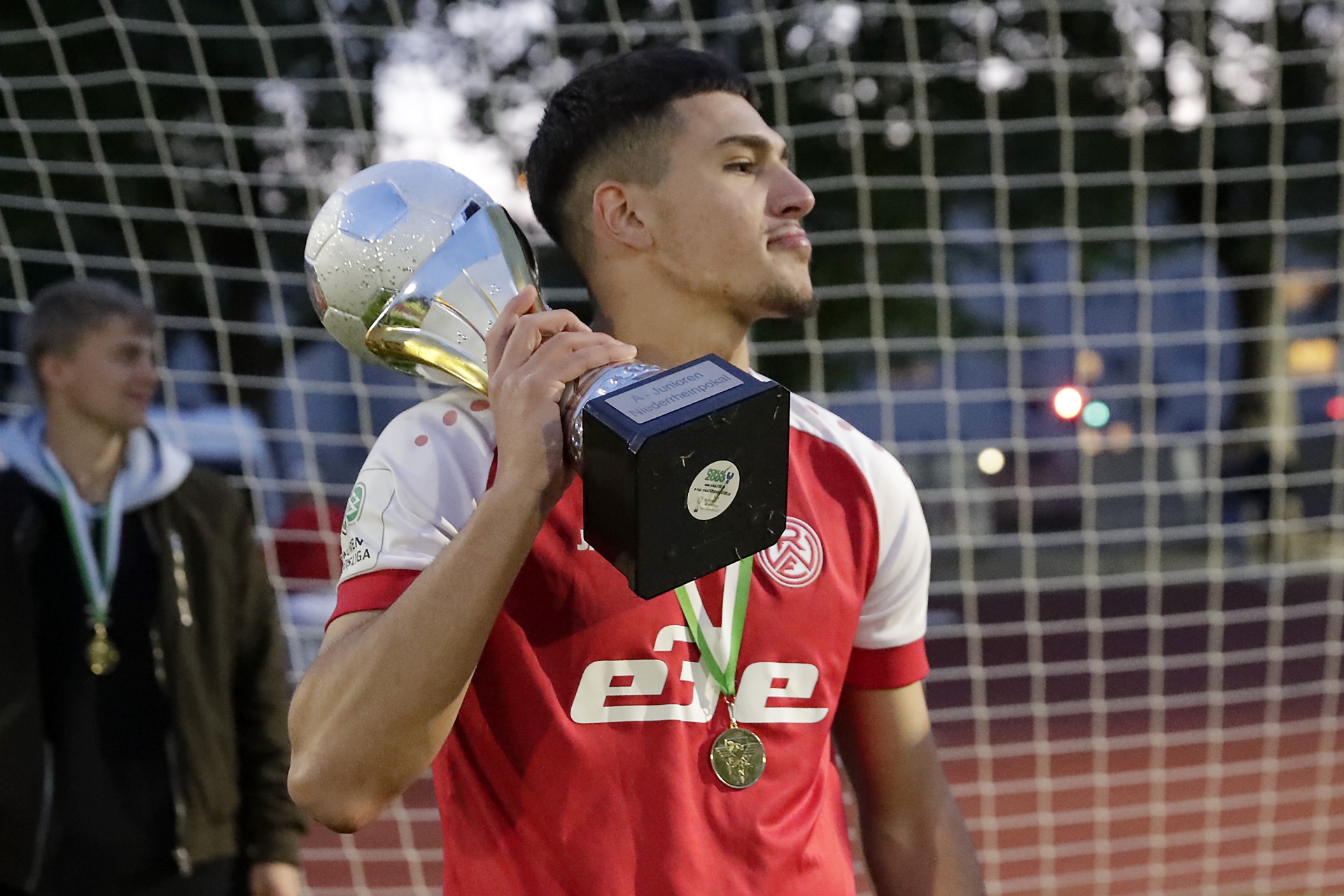 [{"clang_id":1,"value":"Fußballspieler mit Trikot und Medaille hält Pokal über Kopf. Im Hintergrund Netze und verschwommene Personen."}]