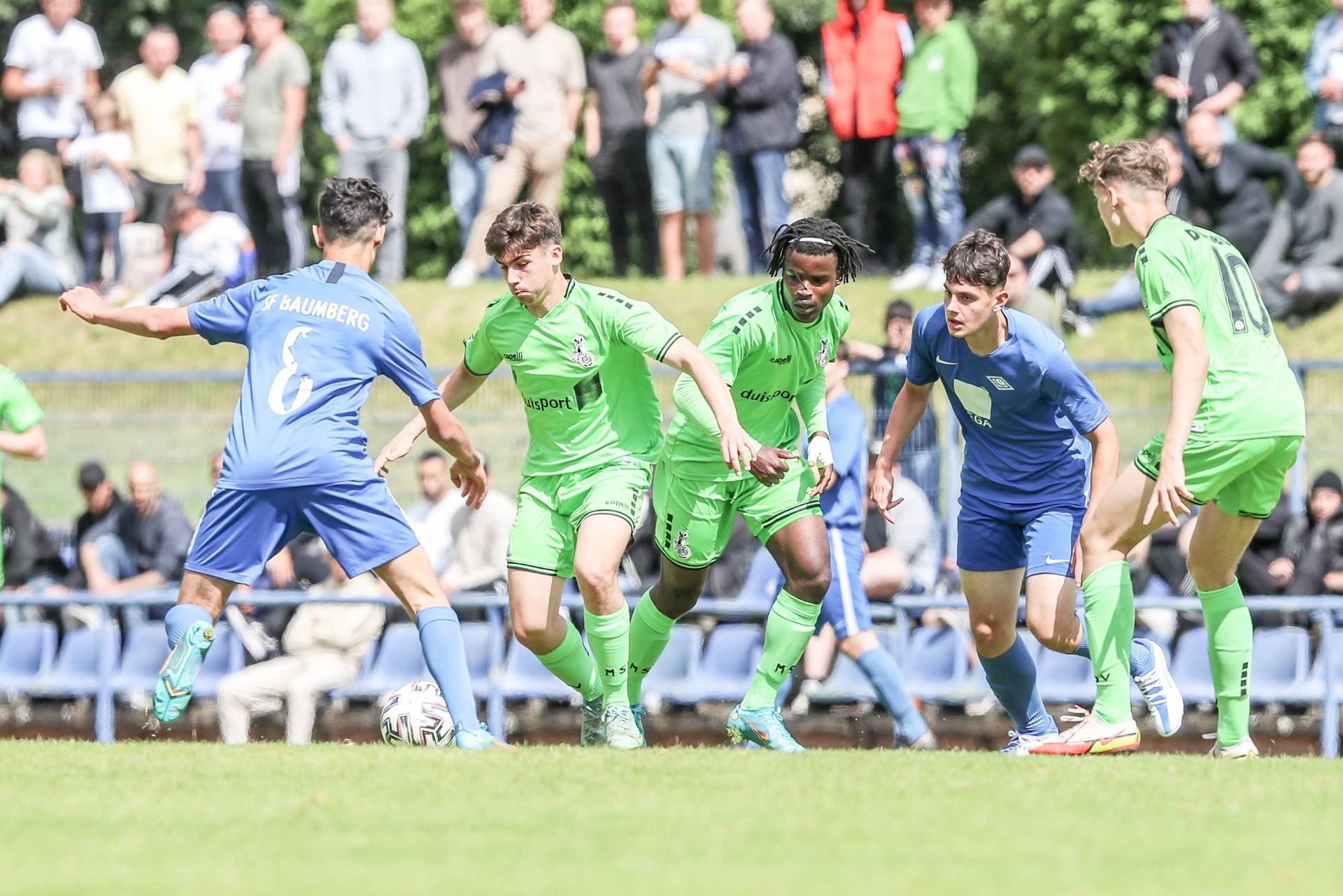 [{"clang_id":1,"value":"Fußballspiel: Spieler in grünen und blauen Trikots kämpfen um den Ball auf einem Rasenplatz. Zuschauer im Hintergrund."}]
