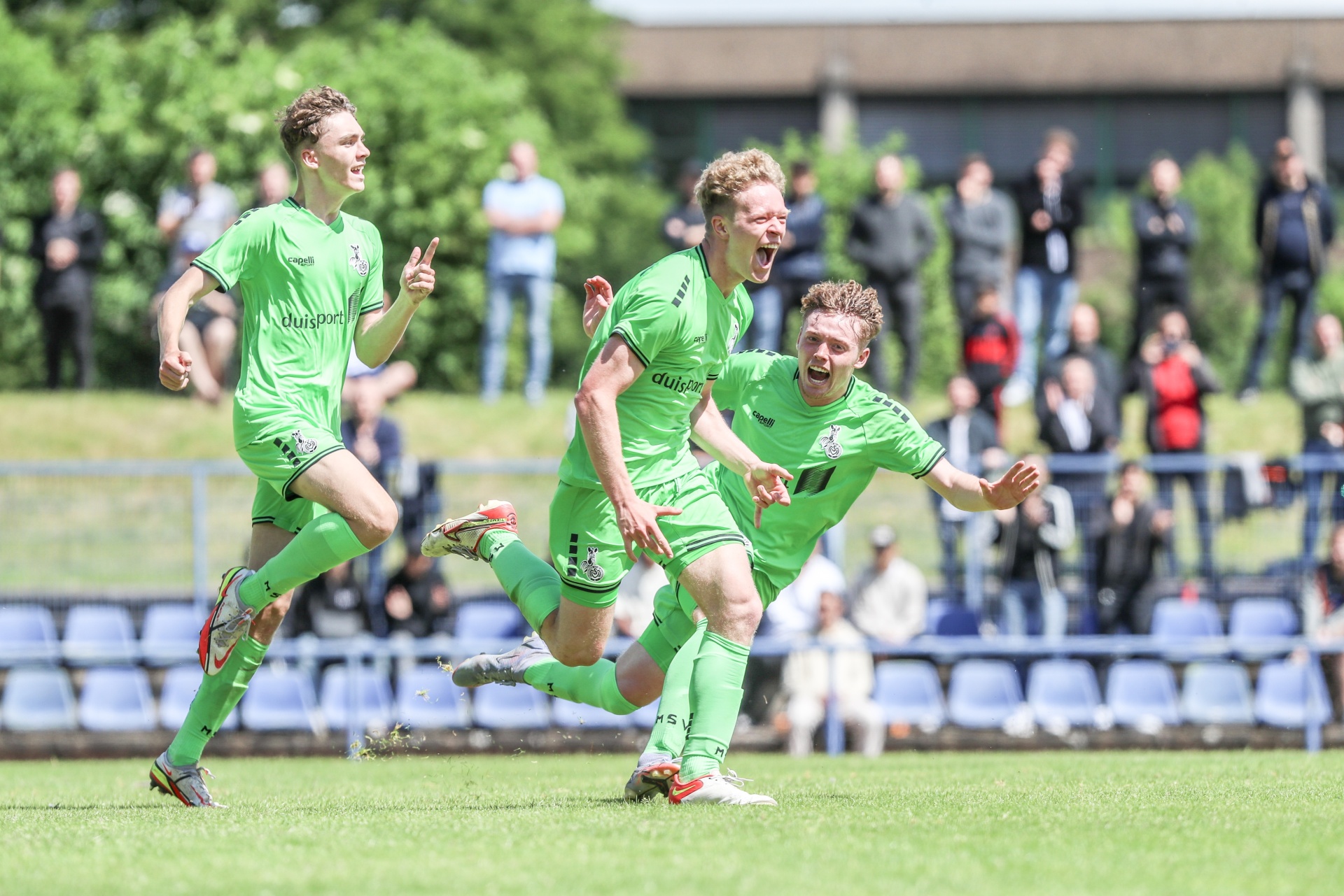 [{"clang_id":1,"value":"Drei Fußballspieler in grünen Trikots jubeln auf einem Rasenplatz, während sie laufen. Im Hintergrund Zuschauer und Bäume."}]