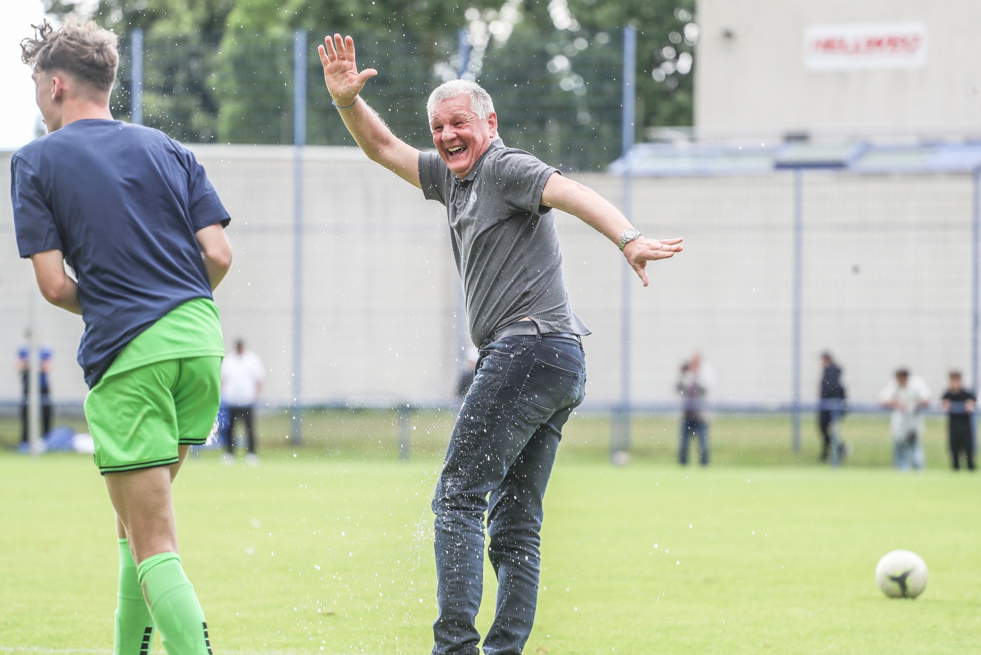 [{"clang_id":1,"value":"Mann wird auf Fußballfeld mit Wasser übergossen, Arme erhoben. Links Spieler in grüner Hose. Hintergrund: Wand und weitere Personen."}]