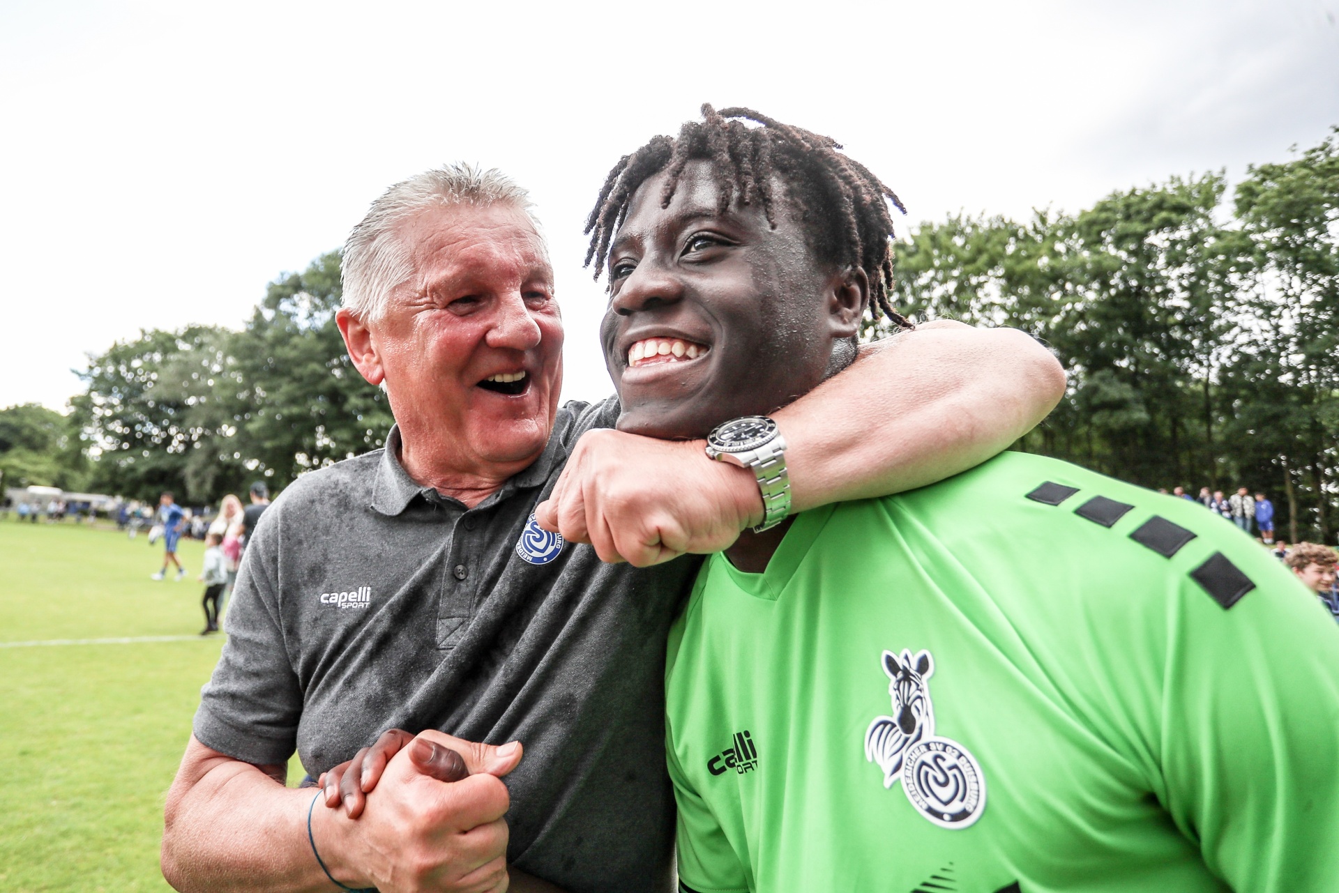 [{"clang_id":1,"value":"Zwei Männer umarmen sich lachend auf einem Fußballplatz. Links ein Mann mit grauem Haar, Polohemd. Rechts ein Mann mit Dreadlocks, grünes Fußballtrikot."}]