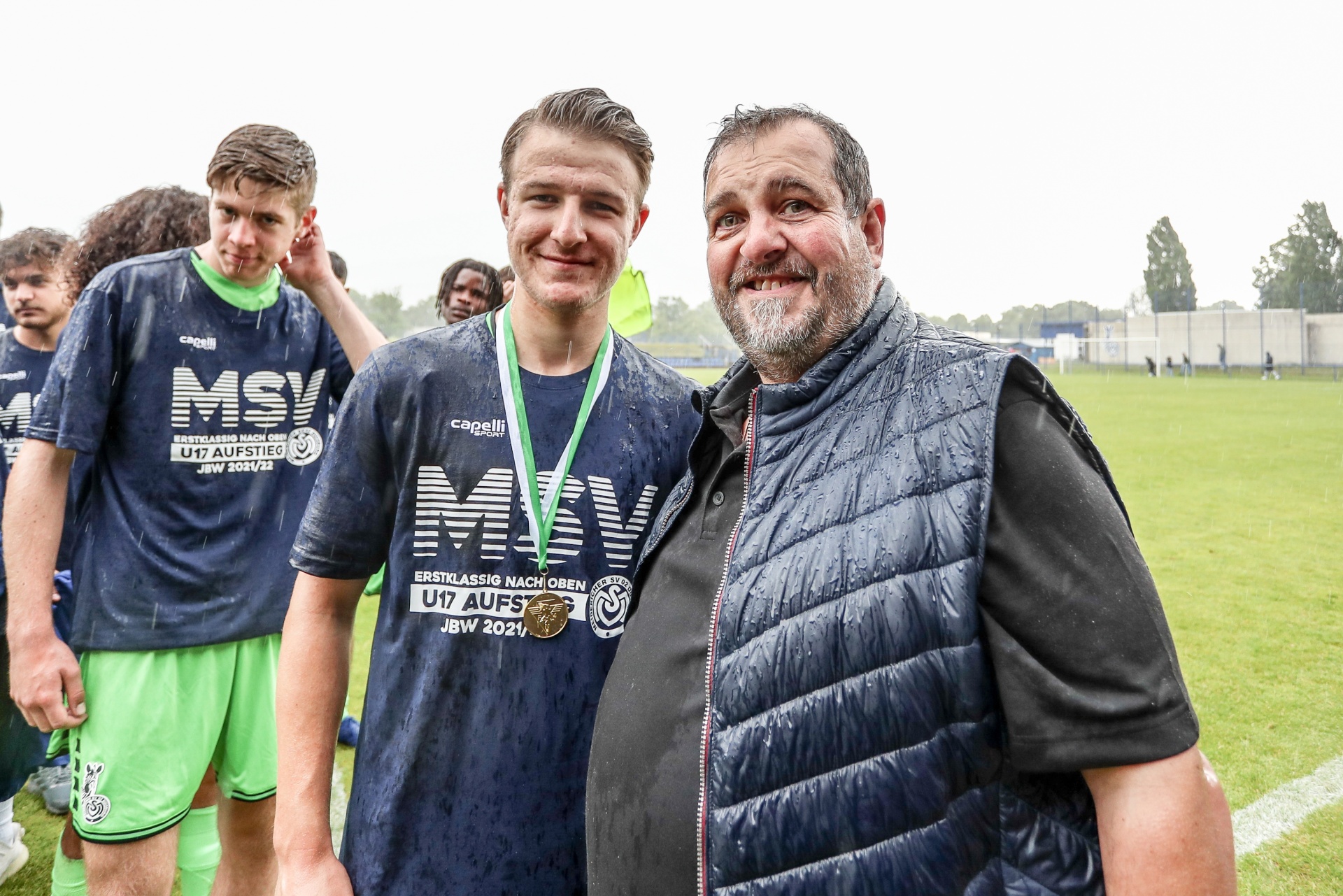 [{"clang_id":1,"value":"Drei Männer auf einem Fußballfeld. Zwei junge Spieler in blau-grünen Trikots, einer mit Medaille, flankiert von einem älteren Mann mit Jacke. Im Hintergrund weitere Spieler."}]