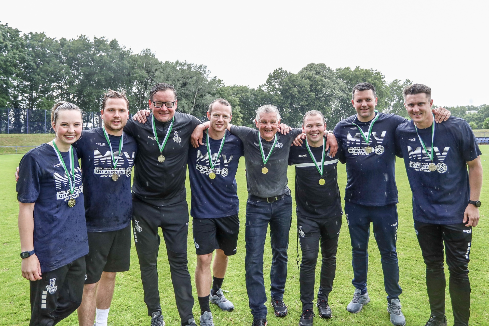 [{"clang_id":1,"value":"Gruppe von acht Personen mit Medaillen um den Hals, stehend auf einem Rasenplatz. Sie tragen dunkle Sportkleidung, ein Mann trägt Jeans und Jacke. Bäume im Hintergrund."}]