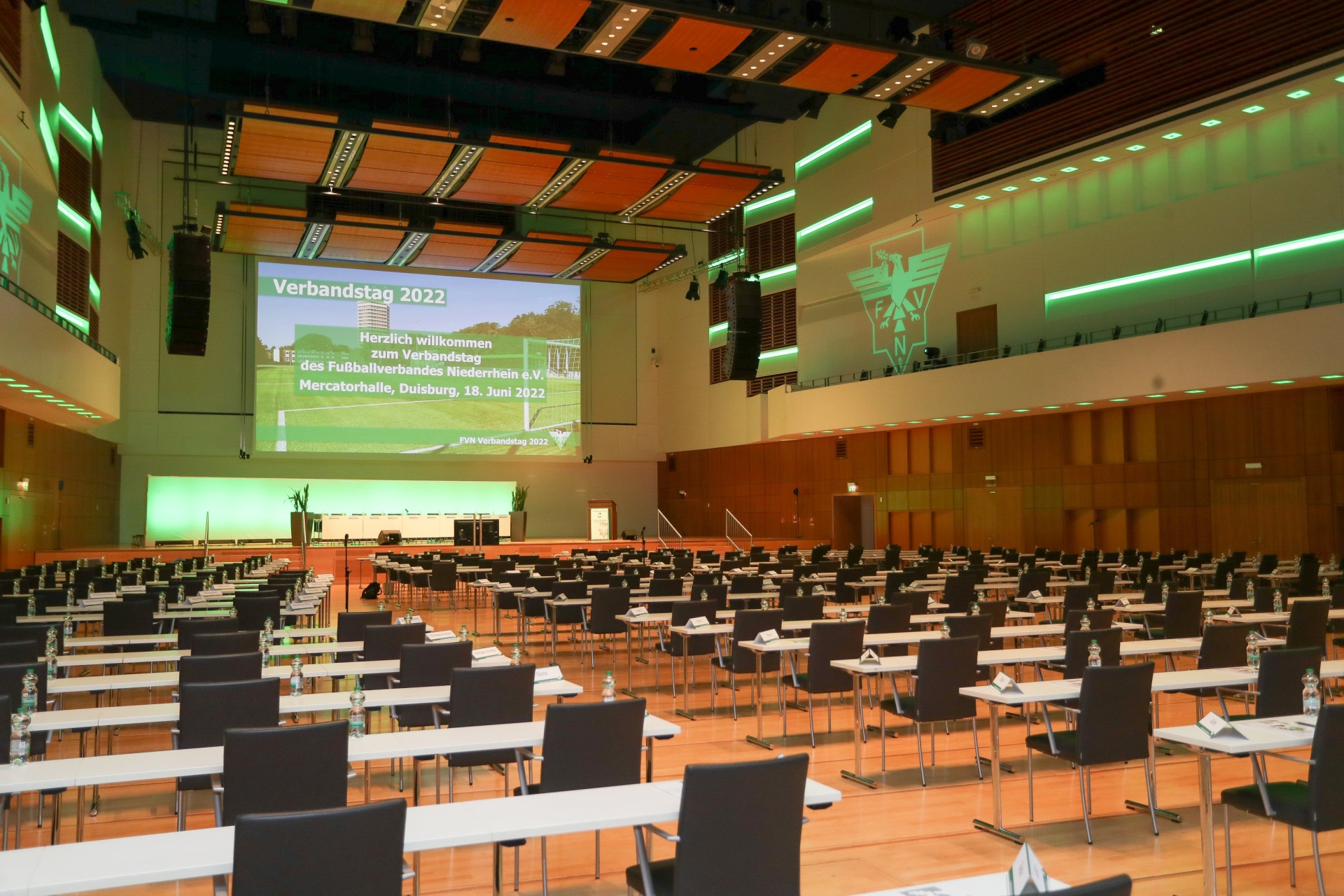 [{"clang_id":1,"value":"Geräumiger Konferenzsaal mit Sitzreihen und Tischen. Auf der Leinwand steht \"Verbandstag 2022\". Grüne Beleuchtung an den Seiten."}]