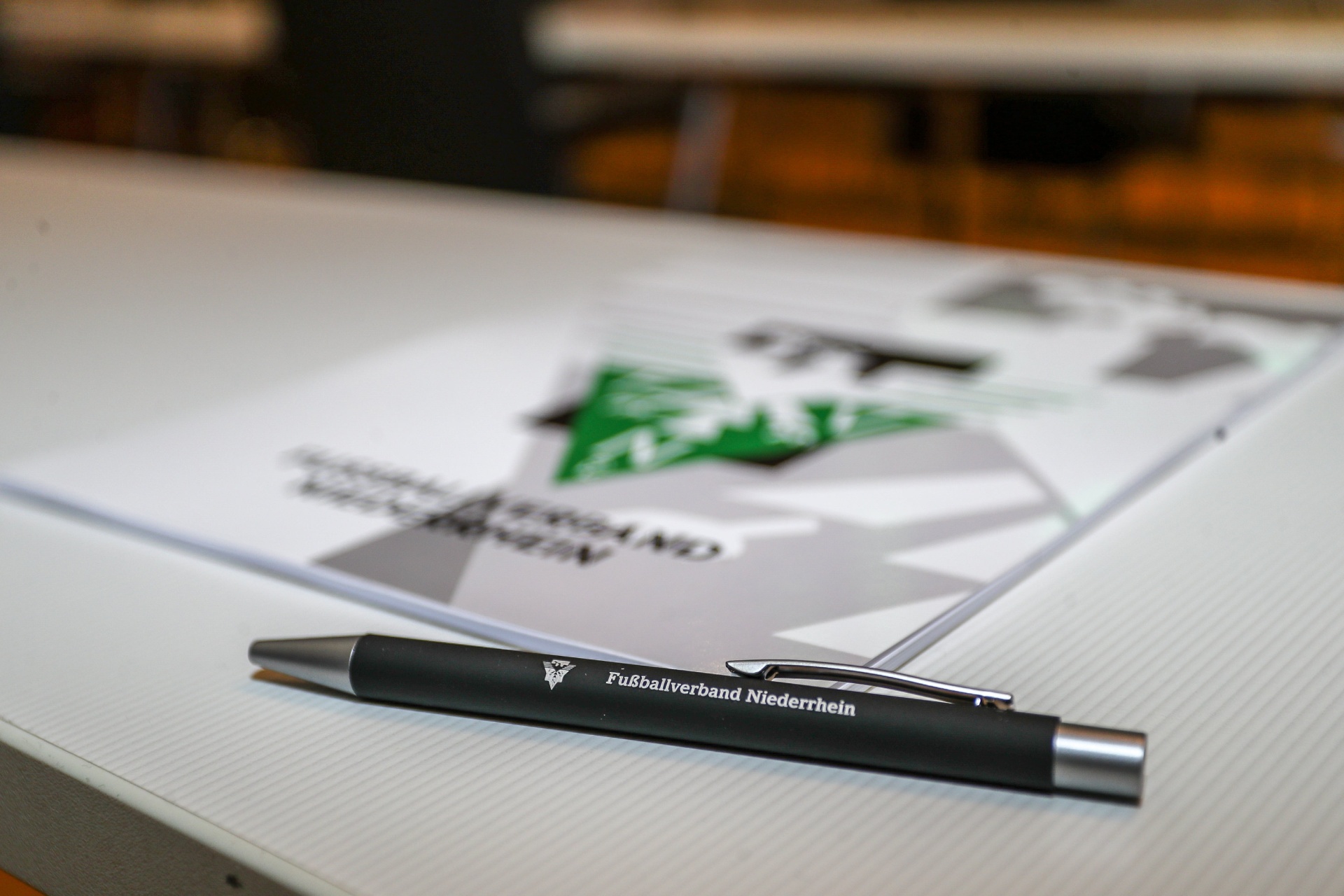 [{"clang_id":1,"value":"Schreibtisch mit Papier und schwarzem Kugelschreiber. Auf dem Papier ist ein Logo mit Fußball-Motiv und dem Text \"Fußballverband Niederhein\" zu sehen."}]
