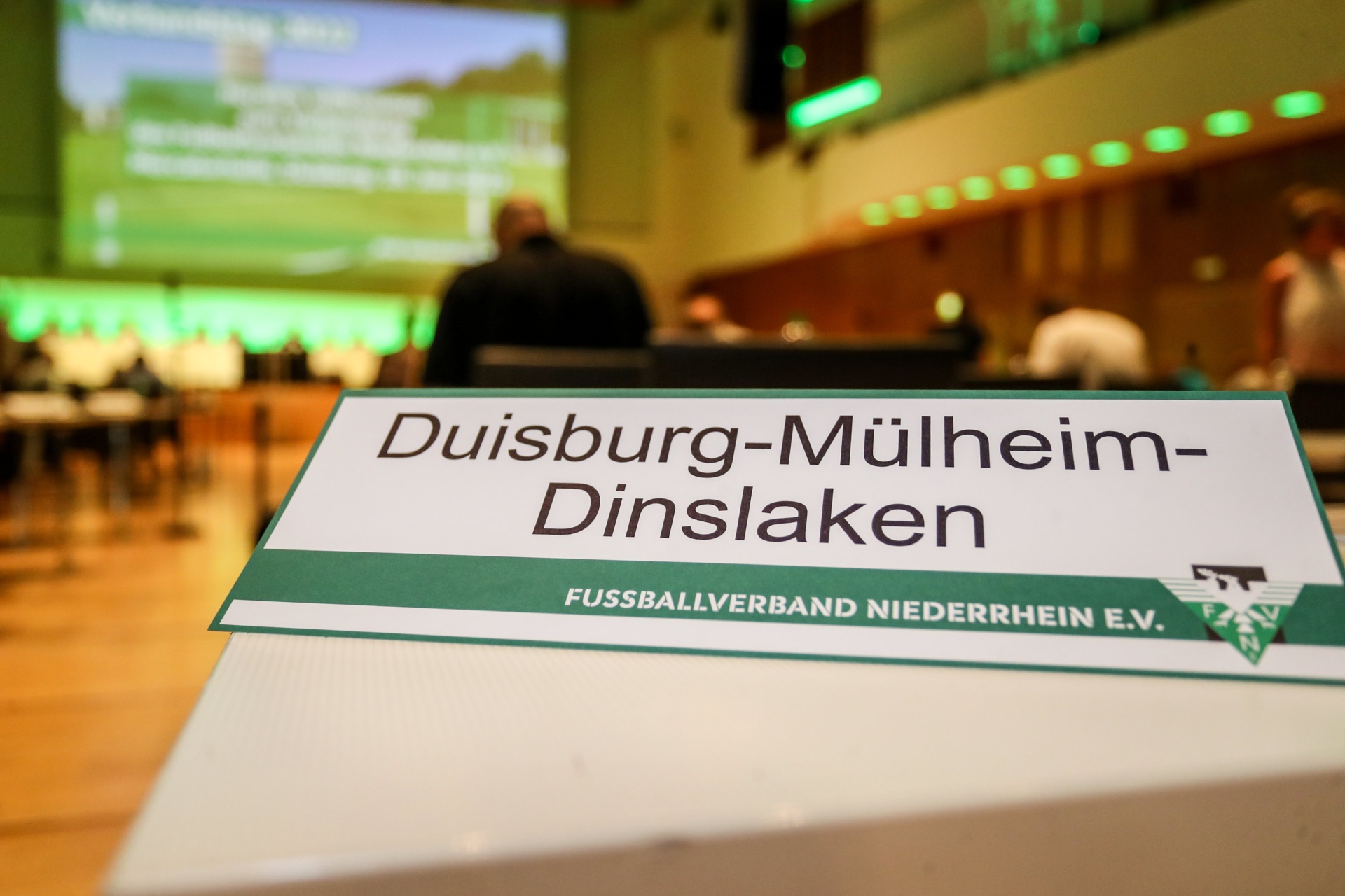 [{"clang_id":1,"value":"Tischkarte mit Aufschrift \"Duisburg-Mülheim-Dinslaken FUSSBALLVERBAND NIEDERRHEIN E.V.\" vor einer Konferenzszene mit Projektion im Hintergrund."}]