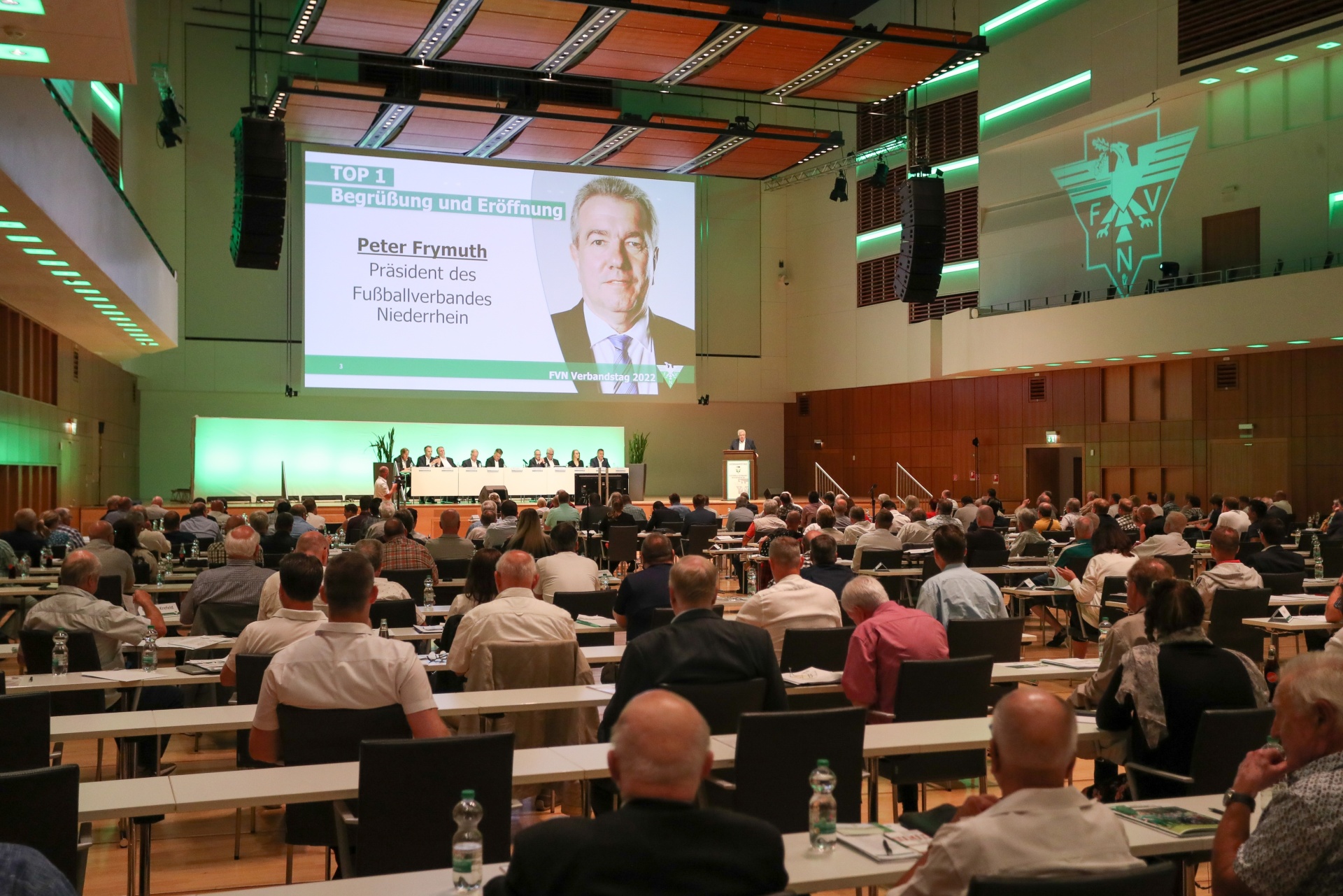 [{"clang_id":1,"value":"Großer Konferenzraum mit Publikum, das einem Vortrag auf einer Leinwand mit dem Porträt von Peter Frymuth, Präsident des Fußballverbandes Niederrhein, lauscht."}]