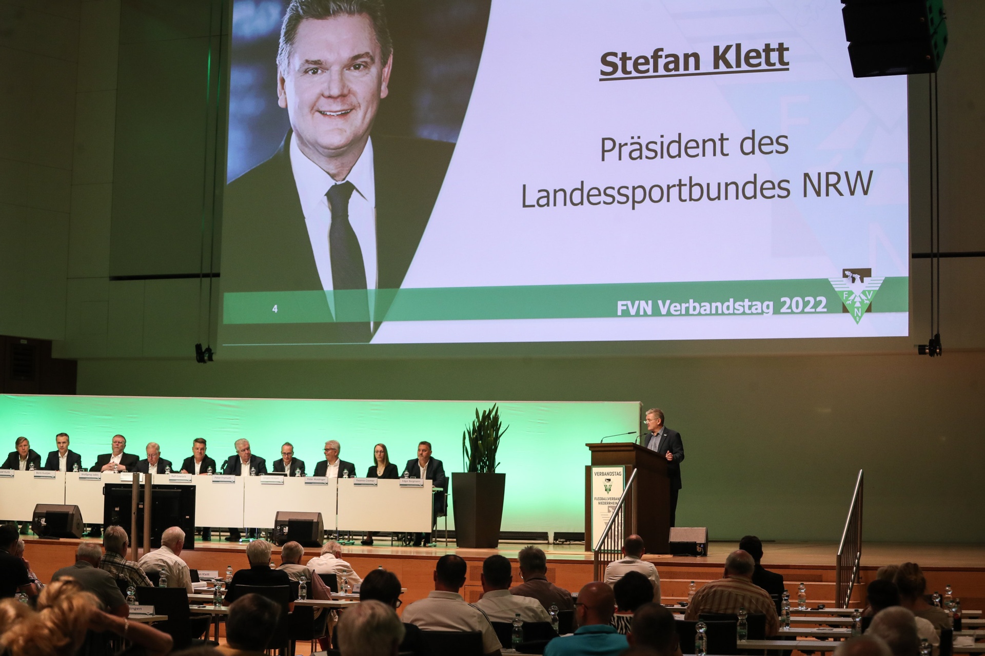 [{"clang_id":1,"value":"Konferenzraum mit Bühne. Auf dem Bildschirm Porträt von Stefan Klett, Präsident des Landessportbundes NRW. Publikum und Podium vor grüner Wand."}]