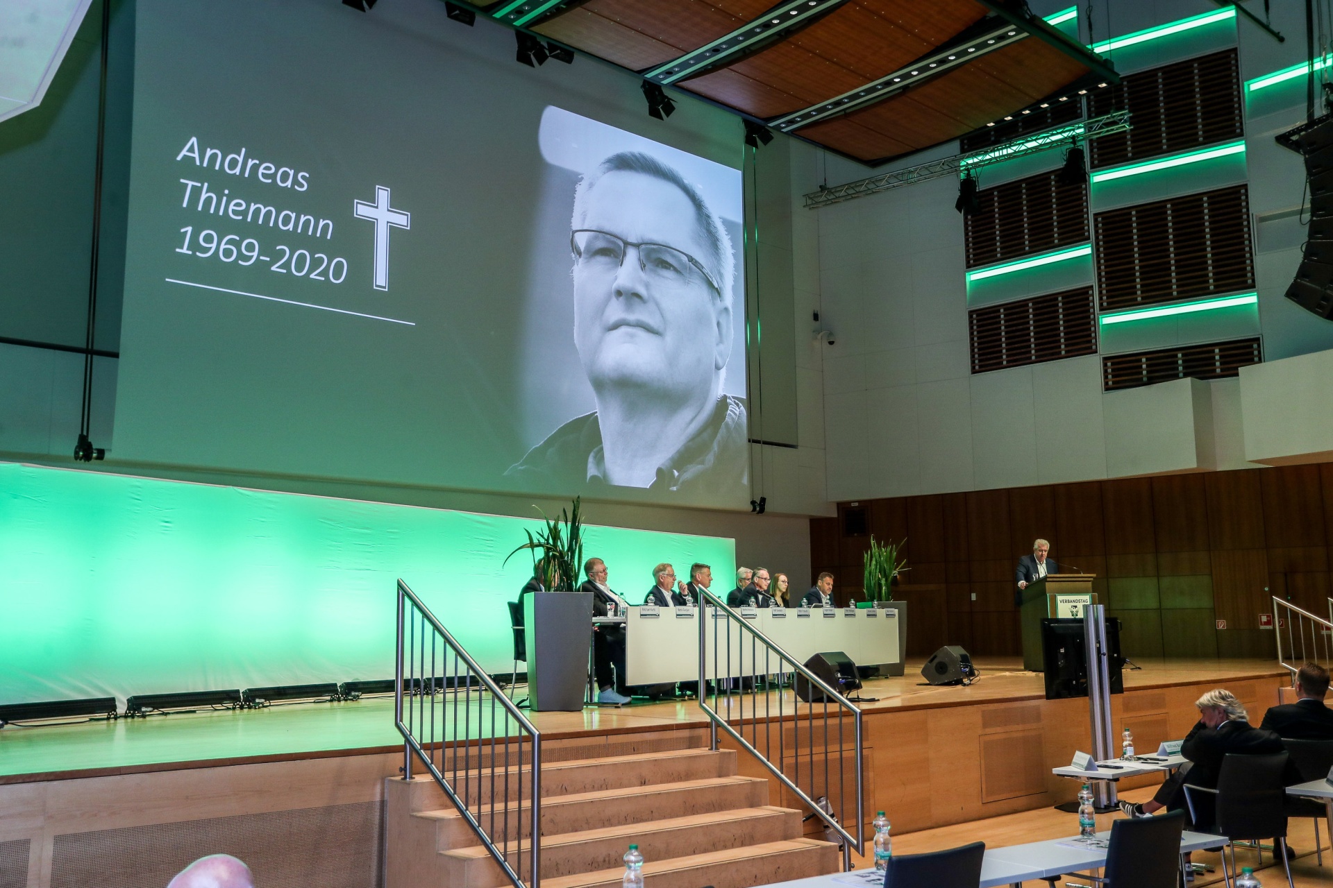 [{"clang_id":1,"value":"Gedenkveranstaltung in einer großen Halle. Im Hintergrund Projektion mit Porträt und Namen \"Andreas Thiemann 1969-2020\". Menschen sitzen an Tischen, Redner am Pult."}]