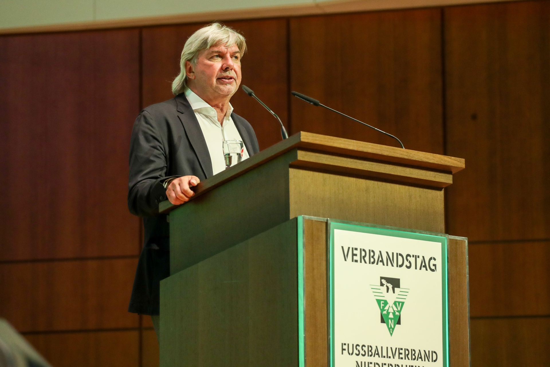 [{"clang_id":1,"value":"Mann mit grauen Haaren spricht an einem Pult. Auf dem Pult steht \"Verbandstag\" und das Logo des Fussballverbands Niederrhein."}]