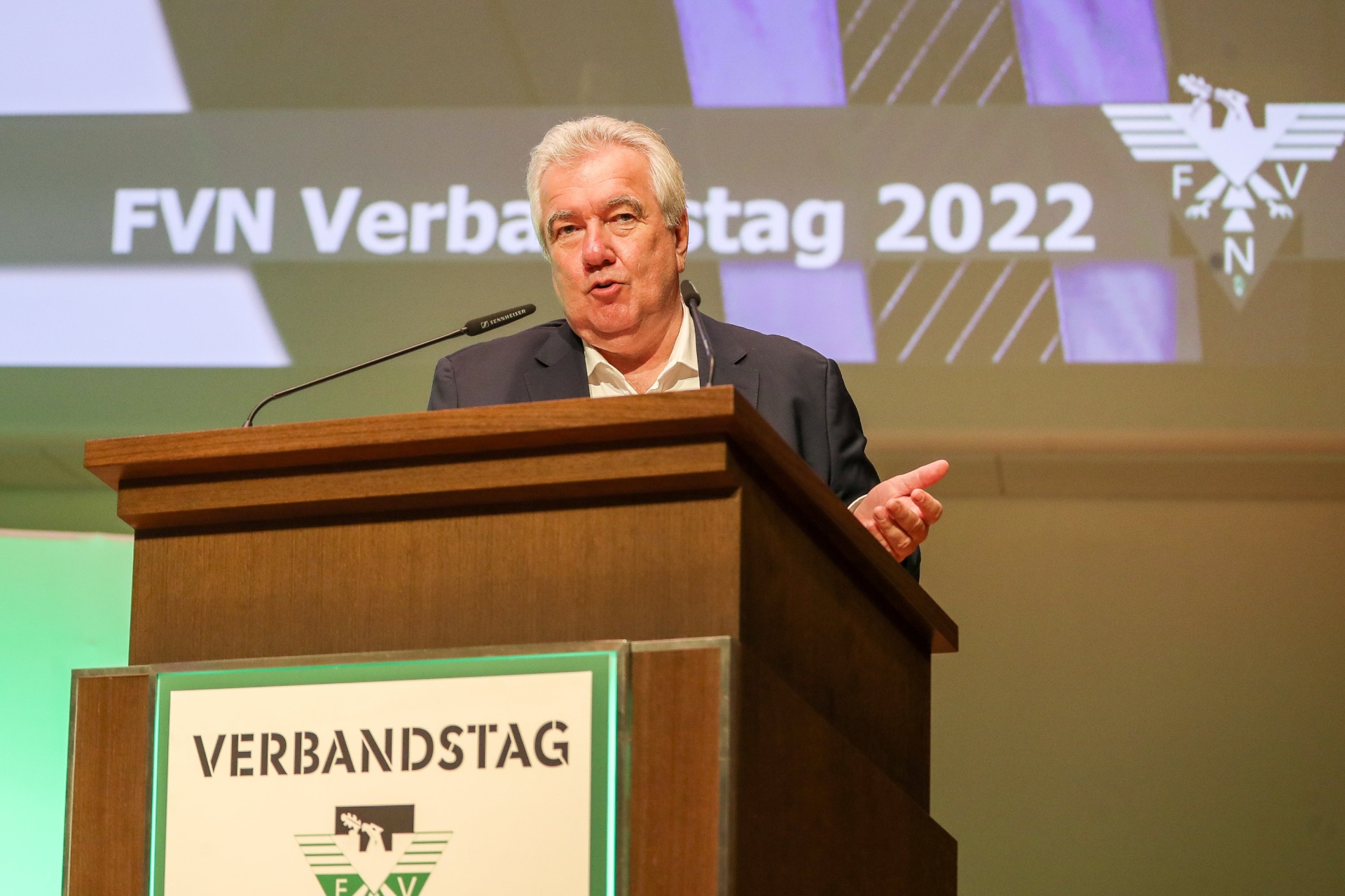 [{"clang_id":1,"value":"Mann am Rednerpult mit Aufschrift \"VERBANDSTAG\" vor Hintergrund mit Schriftzug \"FVN Verbandstag 2022\"."}]