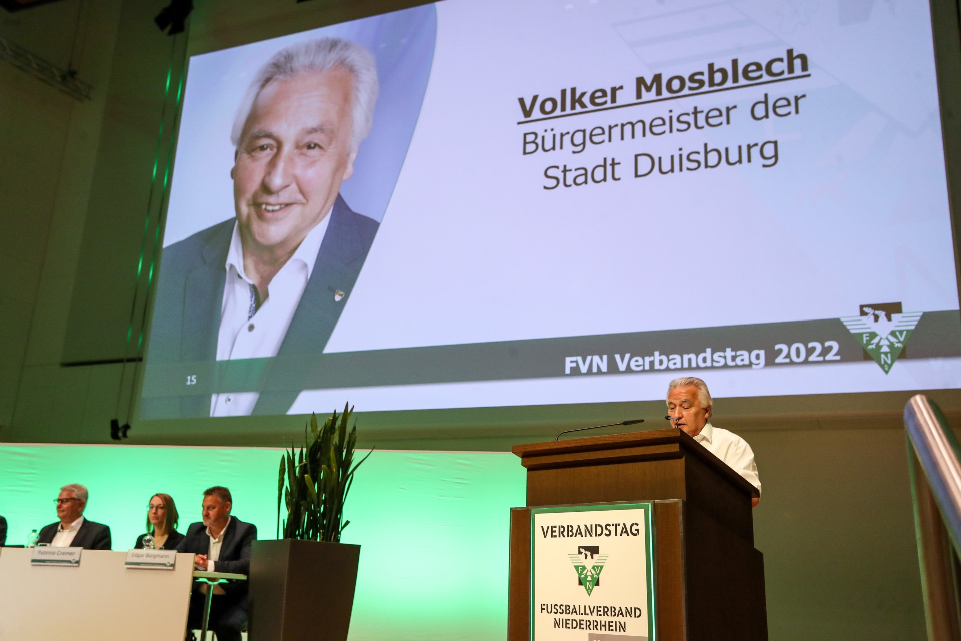 [{"clang_id":1,"value":"Bühne mit Podium. Im Hintergrund Großbildschirm mit Porträt von Volker Mosblech, Bürgermeister von Duisburg. Schriftzug \"FVN Verbandstag 2022\"."}]
