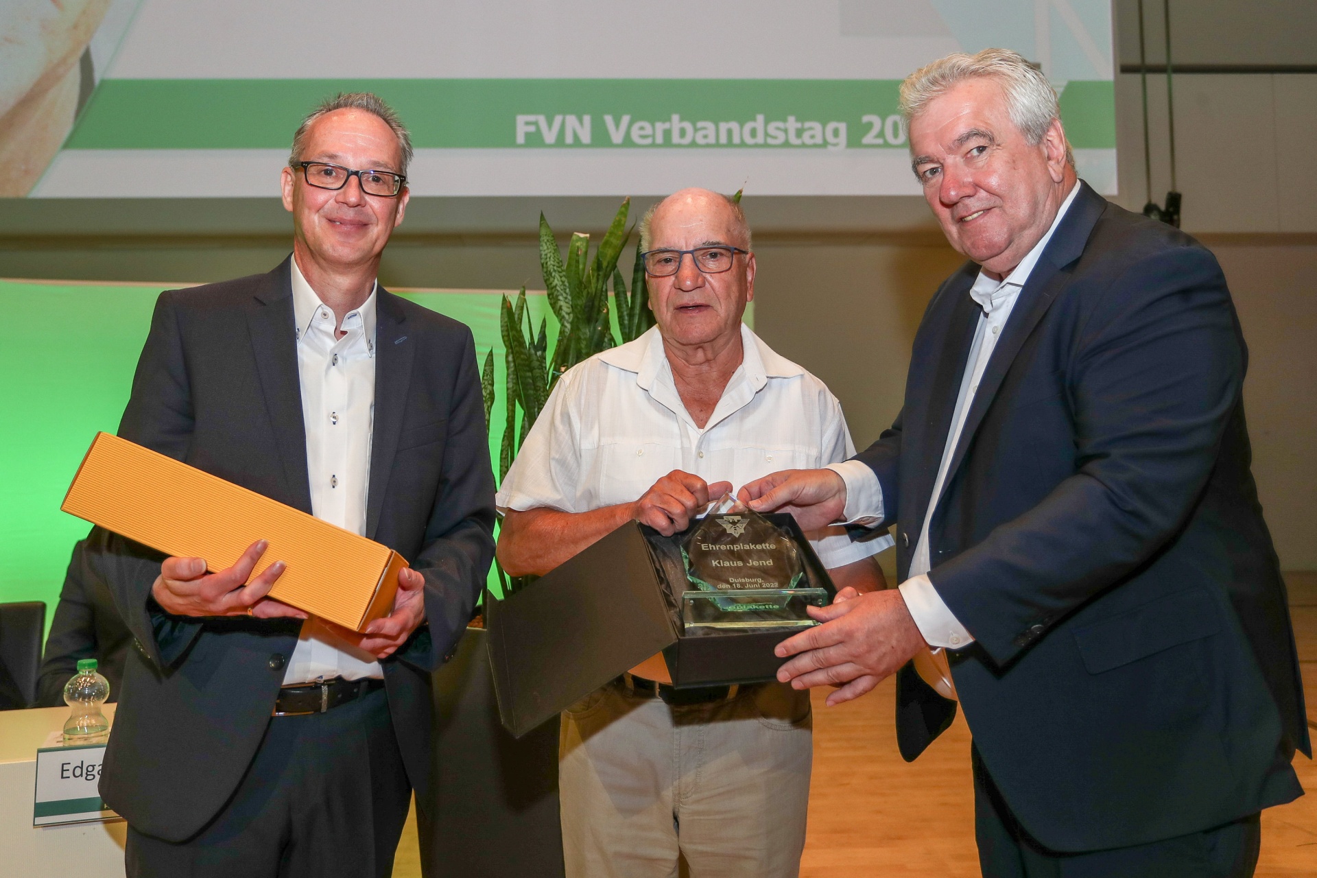 [{"clang_id":1,"value":"Drei Männer auf einer Bühne bei einer Preisverleihung. Der Mann in der Mitte hält eine Auszeichnung, die beiden anderen reichen sie ihm. Im Hintergrund ein Banner mit \"FVN Verbandstag 20..\"."}]