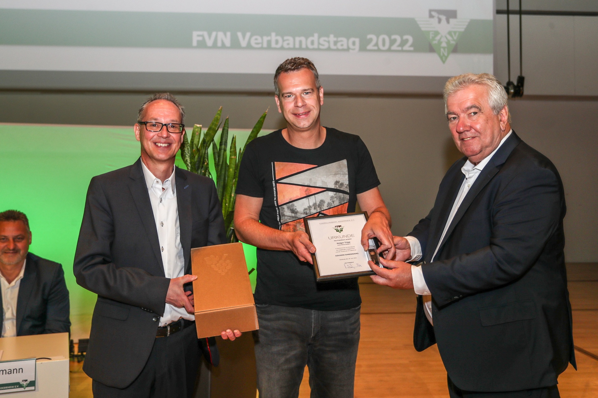 [{"clang_id":1,"value":"Drei Männer auf einer Bühne. Mitte ein Mann mit Urkunde und Geschenk, links und rechts je ein Mann im Anzug. Im Hintergrund Schriftzug \"FVN Verbandstag 2022\"."}]