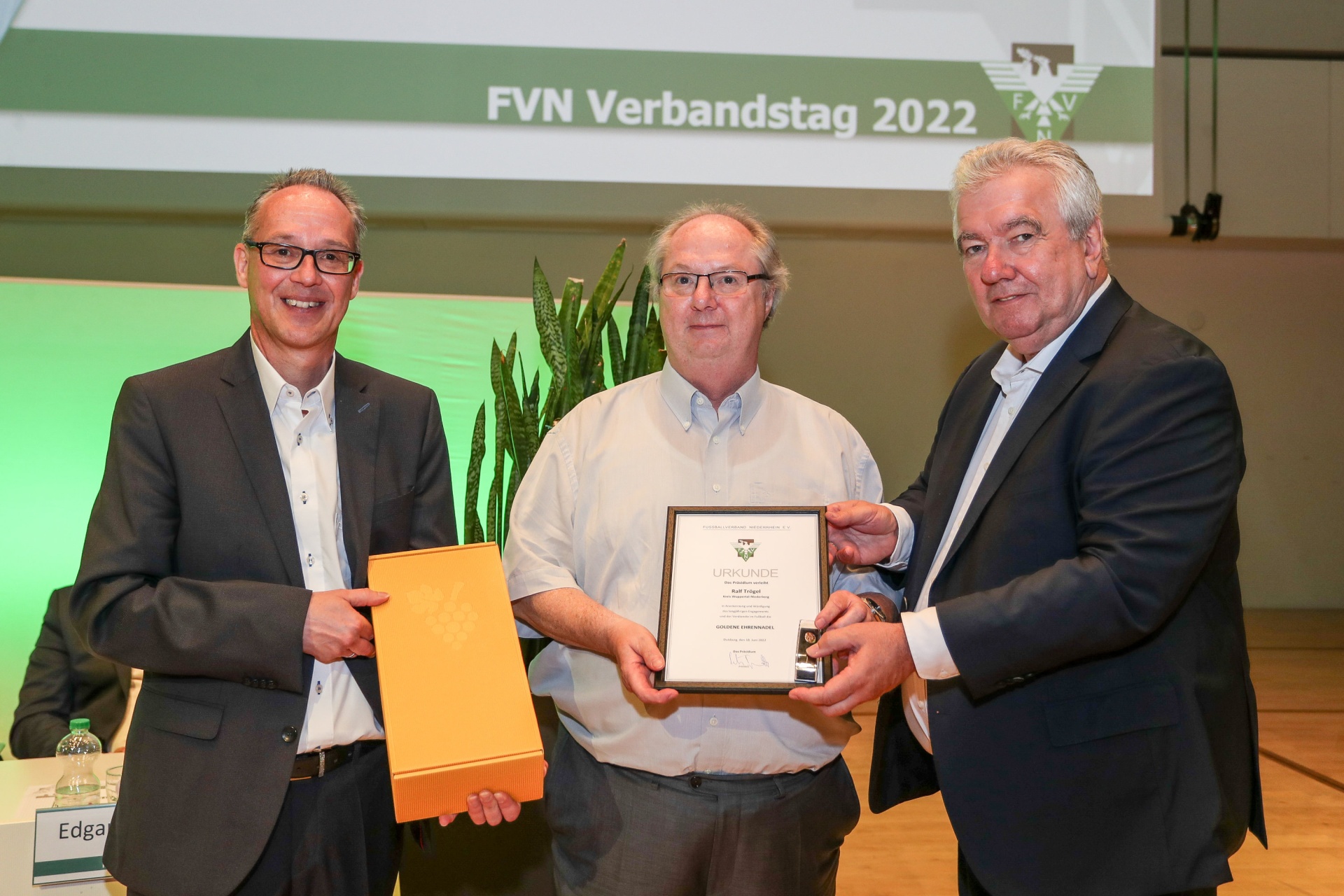 [{"clang_id":1,"value":"Drei Männer bei einer Preisverleihung. Links und rechts überreichen sie dem Mann in der Mitte Urkunden und eine goldfarbene Plakette. Im Hintergrund ein Banner mit dem Text \"FVN Verbandstag 2022\"."}]