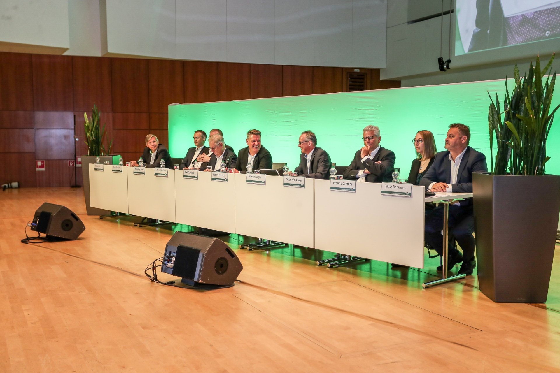 [{"clang_id":1,"value":"Zehn Personen sitzen an einem Tisch auf einer Bühne vor einem grünen Hintergrund. Mikrofone und Namensschilder sind sichtbar. Ein großer Raum mit Publikum ist angedeutet."}]