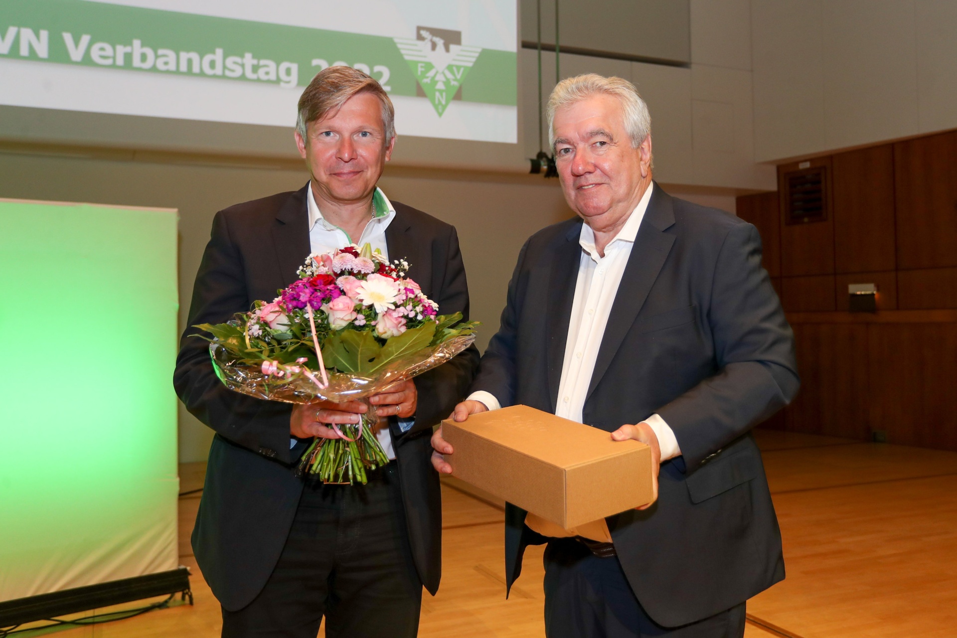 [{"clang_id":1,"value":"Zwei Männer stehen auf einer Bühne. Links hält einer Blumen, rechts übergibt der andere eine Geschenkbox. Im Hintergrund ein Banner mit Schriftzug \"VN Verbandstag 2022\"."}]