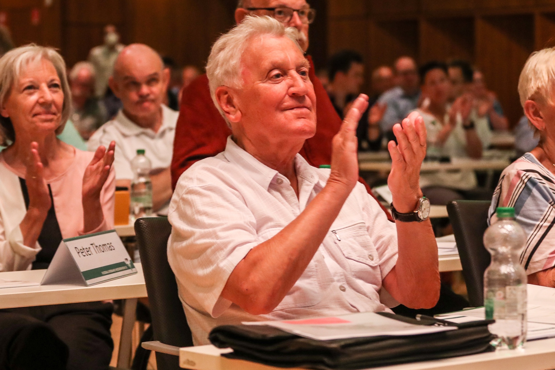 [{"clang_id":1,"value":"Zuschauer in einem Raum applaudieren. Im Vordergrund ein Mann mit grauen Haaren und Hemd. Vor ihm Namensschild \"Peter Thomas\"."}]