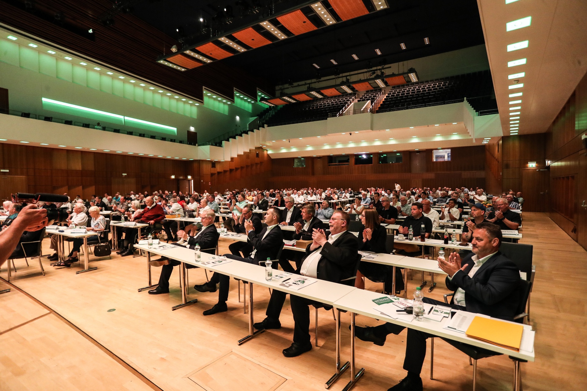 [{"clang_id":1,"value":"Großer Konferenzsaal mit zahlreichen Zuschauern auf Stühlen vor Tischen. Im Vordergrund sitzen und klatschen mehrere Männer in Anzügen."}]